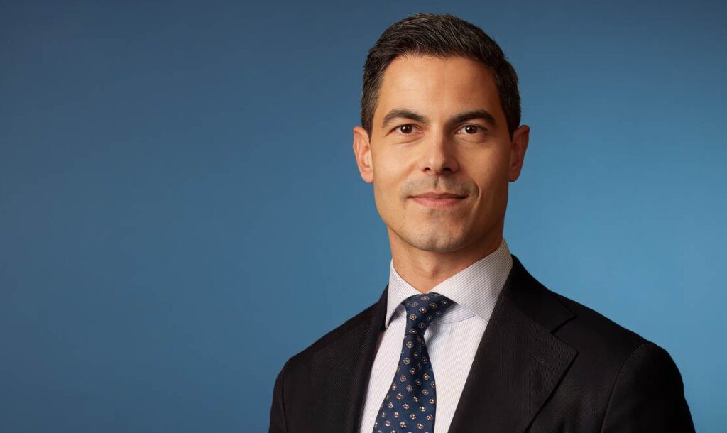 Professional head-and-shoulders portrait of a man in a dark suit, light shirt, and patterned tie against a blue backdrop.