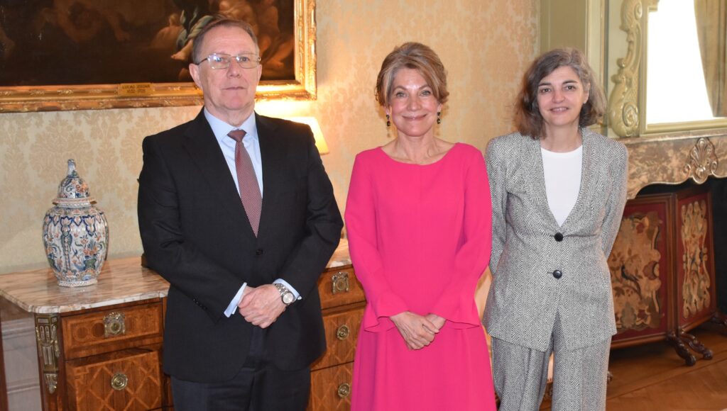 Three formally dressed individuals posing for a photo in an ornate room, with a large painting, gold accents, and a decorative vase on a marble table nearby (man left, woman in pink center, woman in gray suit right).