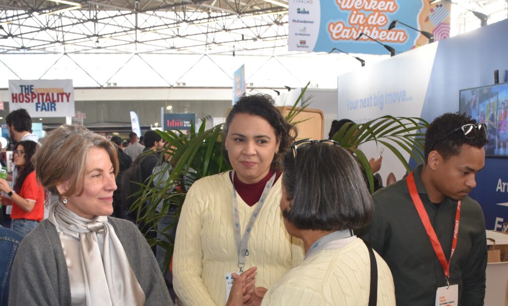 Three women wearing lanyards converse at a busy hospitality fair, with banners and booths in the background.