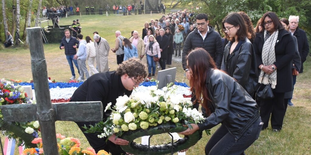 Mourners lay a large floral wreath at a cemetery grave during a memorial service, with a crowd gathered in the background.