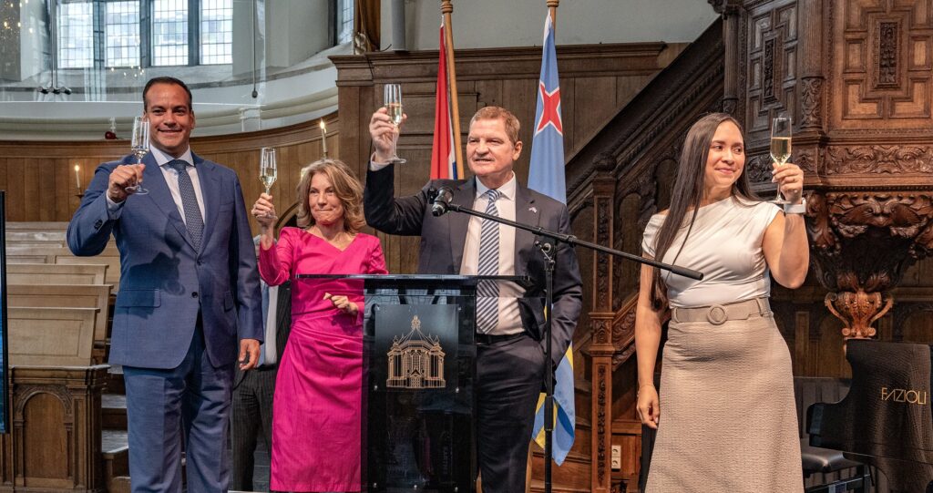 Four adults in formal attire toast with champagne around a podium during a ceremony in a grand wood-paneled hall with flags in the background, smiling earnestly on their faces to the camera.