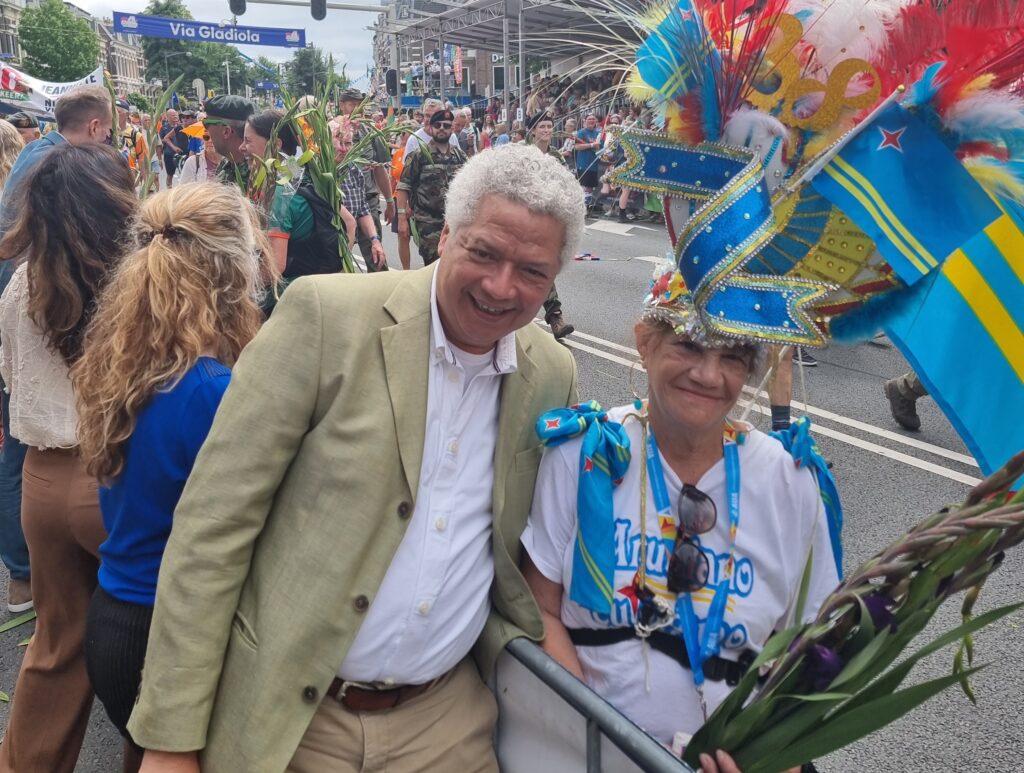Smiling man in a beige suit and a festive crowd pose at a colorful street parade with a banner overhead reading 'Via Gladiola'.