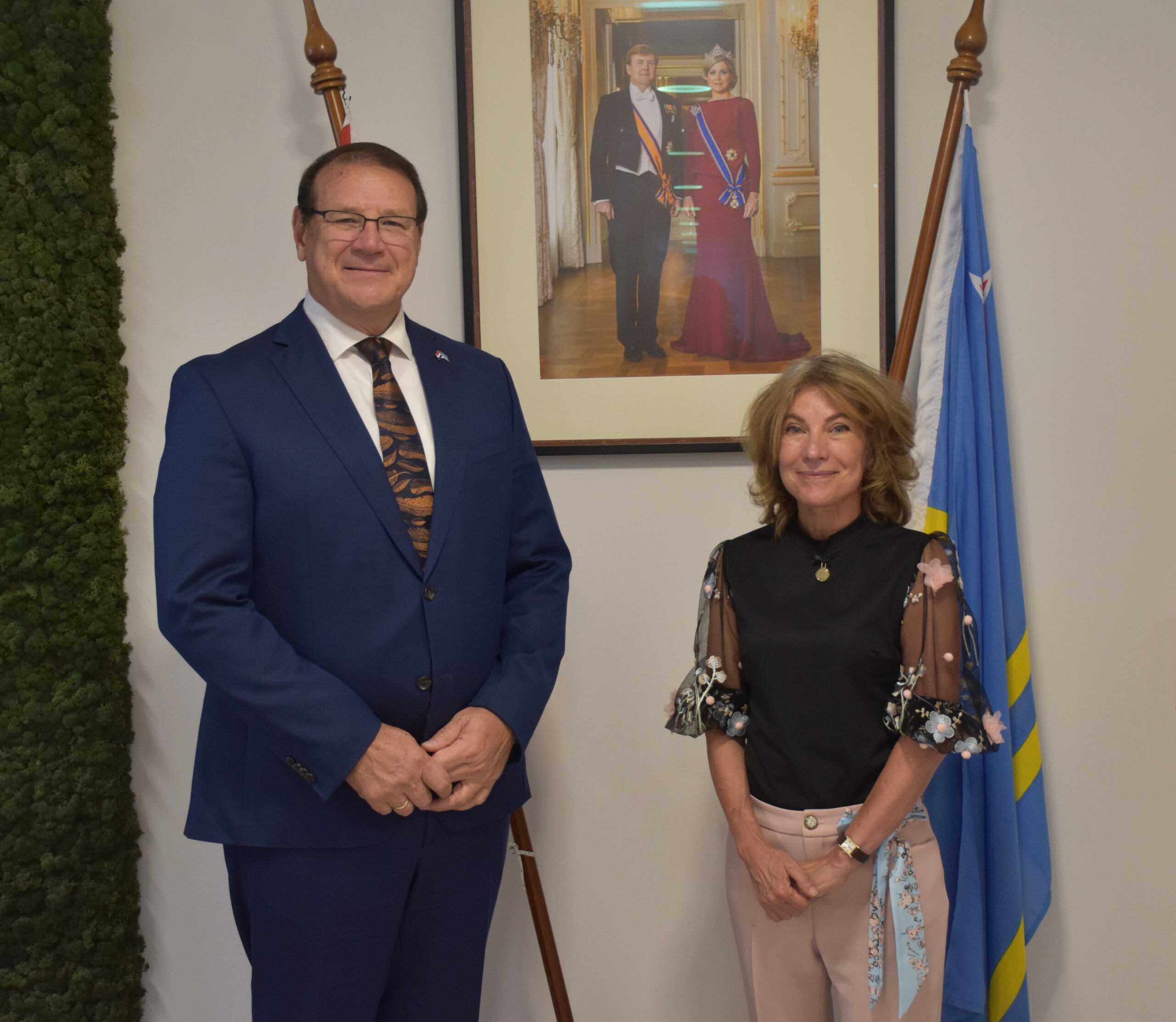 Two adults pose indoors in front of a framed portrait of a royal couple; a blue and yellow flag is on the right.