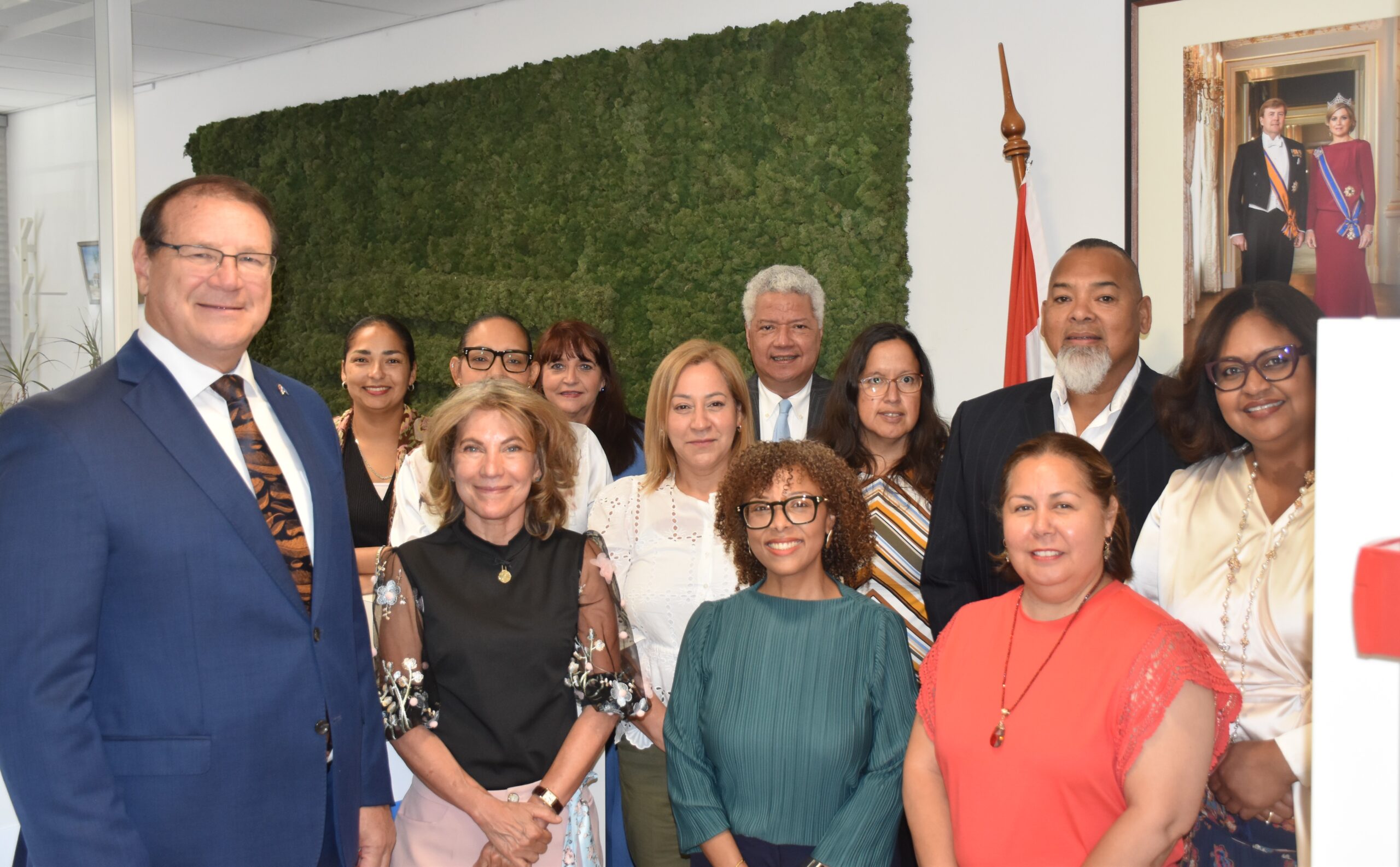 Group of diverse adults in formal attire posing for a group photo in an office lobby with a green wall and flags visible in the background.