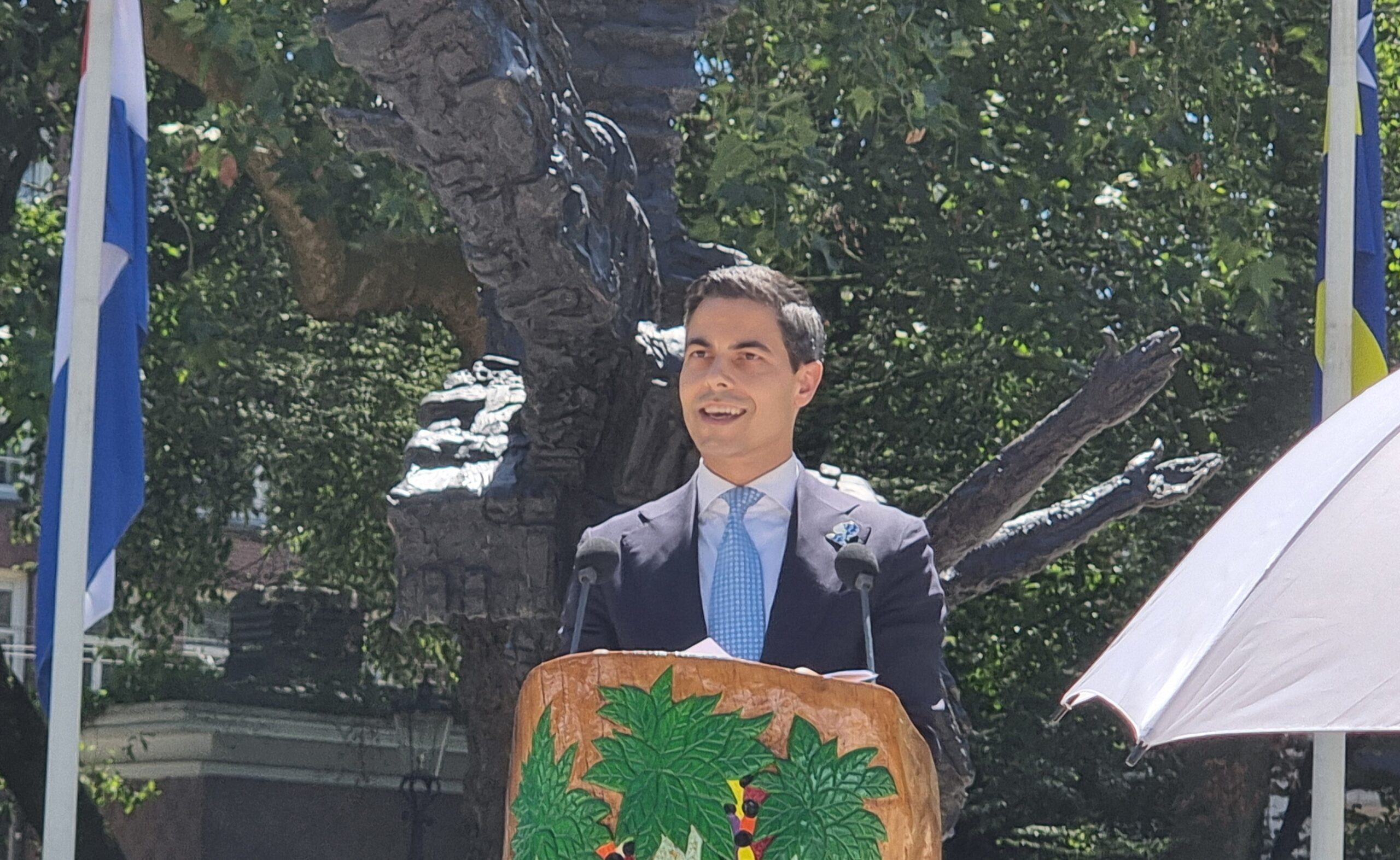Man in a suit delivers a speech at an outdoor ceremony, standing behind a leaf-patterned podium with flags in the background.