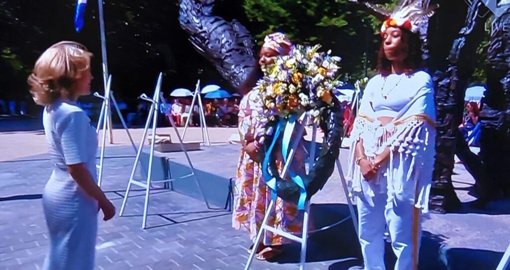 Woman in a white dress stands facing two performers in ceremonial attire outdoors, one with a flower garland and bright headdress and another in white with fringe sleeves, near white frames and chairs.