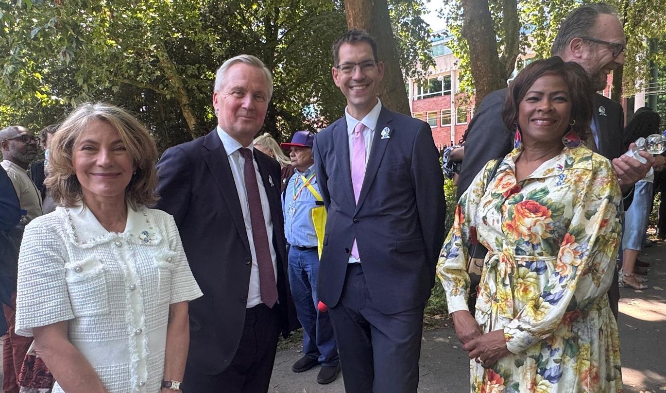 Four professionally dressed people posing outdoors at a daytime event, smiling with trees and a brick building in the background.