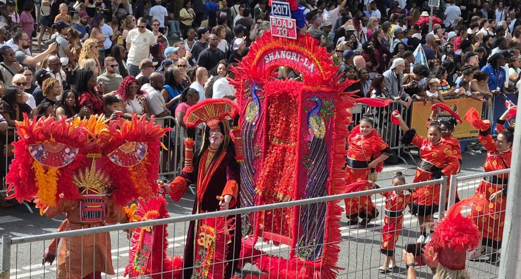 Vibrant dragon parade with performers in red and orange costumes marching along a street as spectators look on from behind barriers.
