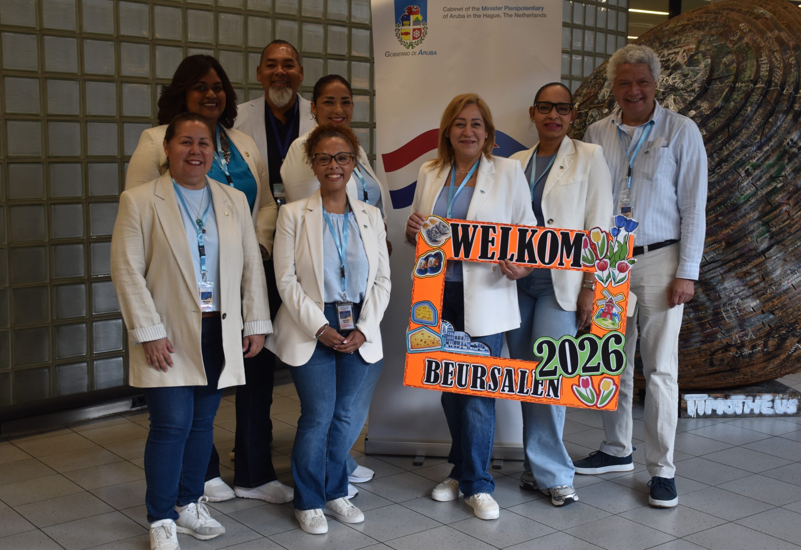 Group of diverse professionals in white coats posing indoors with a bright colorful 'WELCOME 2026' sign behind them and a banner in the background.