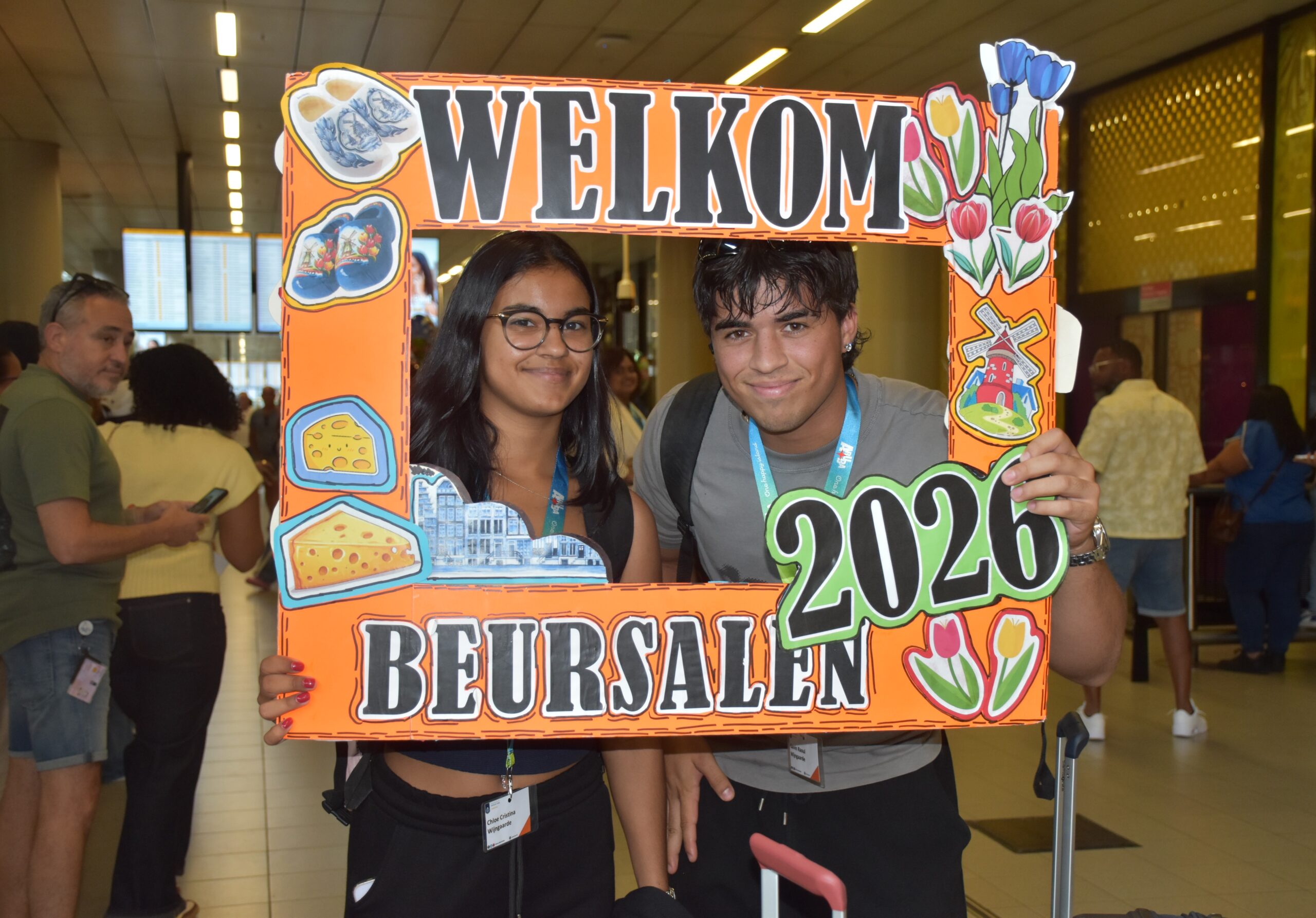 Two young people smile behind a bright orange photo frame reading 'WELCOME 2026' adorned with flowers and cheese icons at an indoor event.