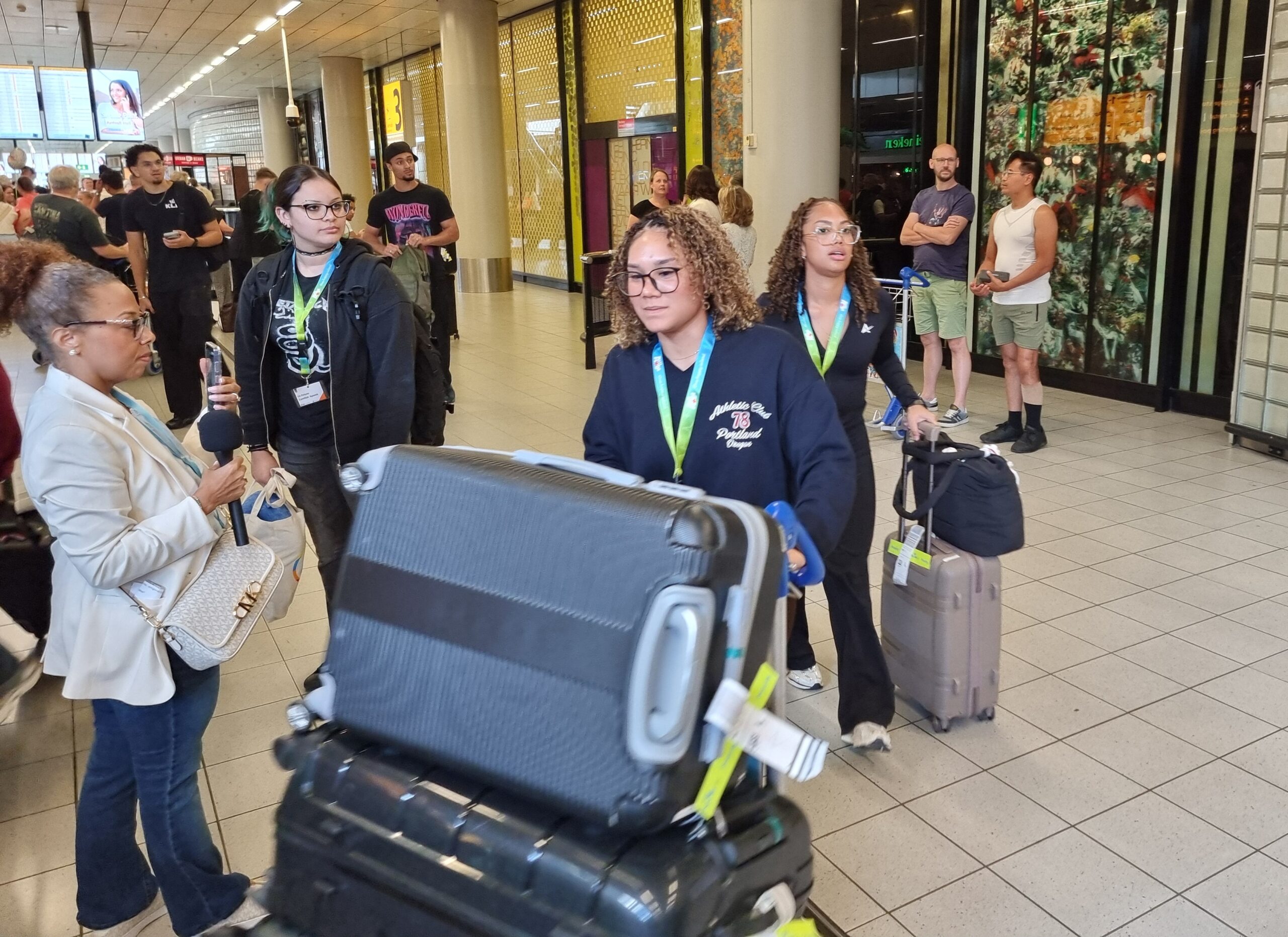 Reporter interviewing a traveler with a microphone while others queue with suitcases in a busy airport terminal.