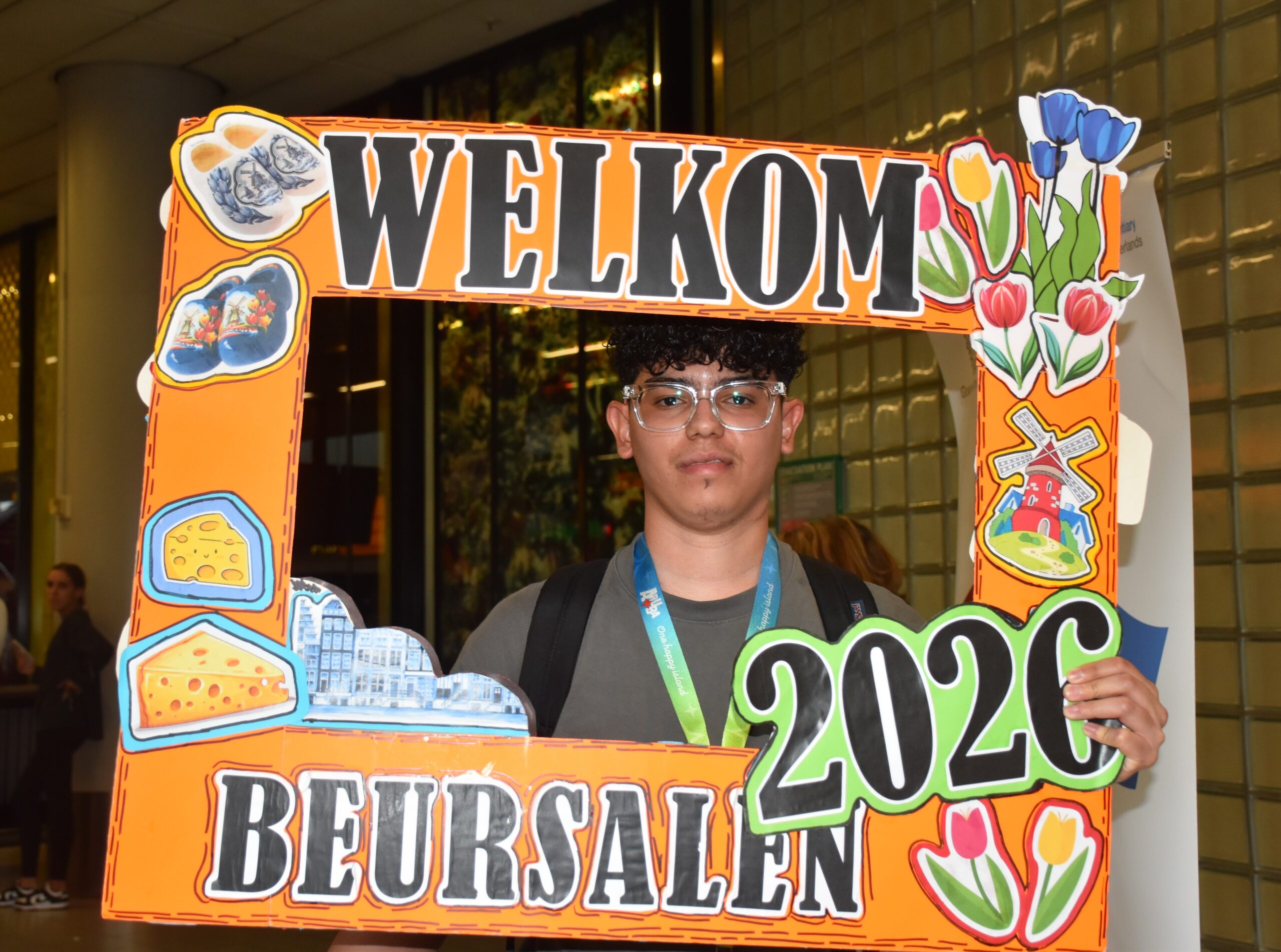 Young man with glasses and a lanyard holds a large orange decorative 'WELCOME' sign frame indoors, smiling at the camera.
