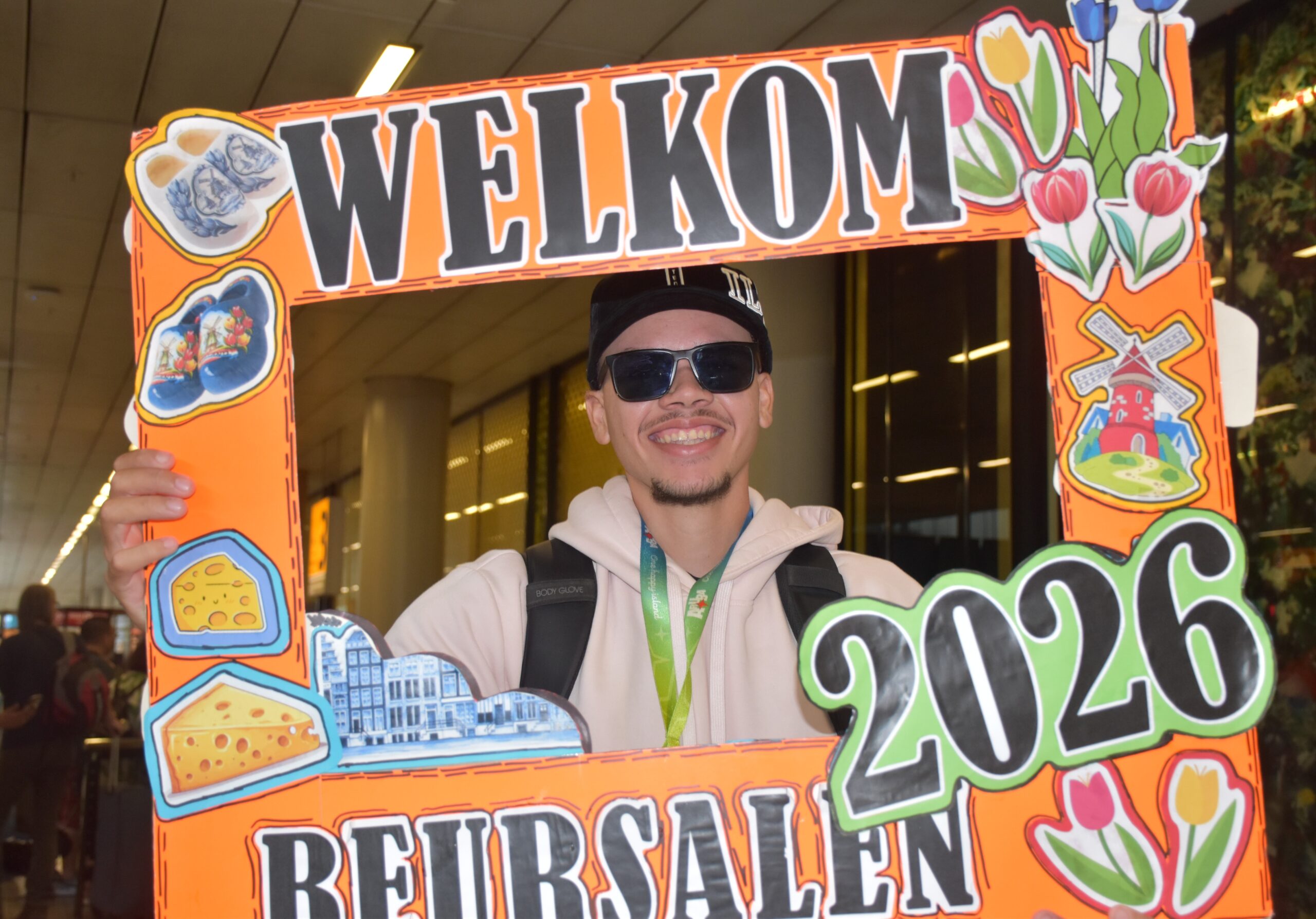 Smiling man in sunglasses and a backpack holds a large orange welcome-frame sign with plant and food illustrations that reads 'WELCOME'.
