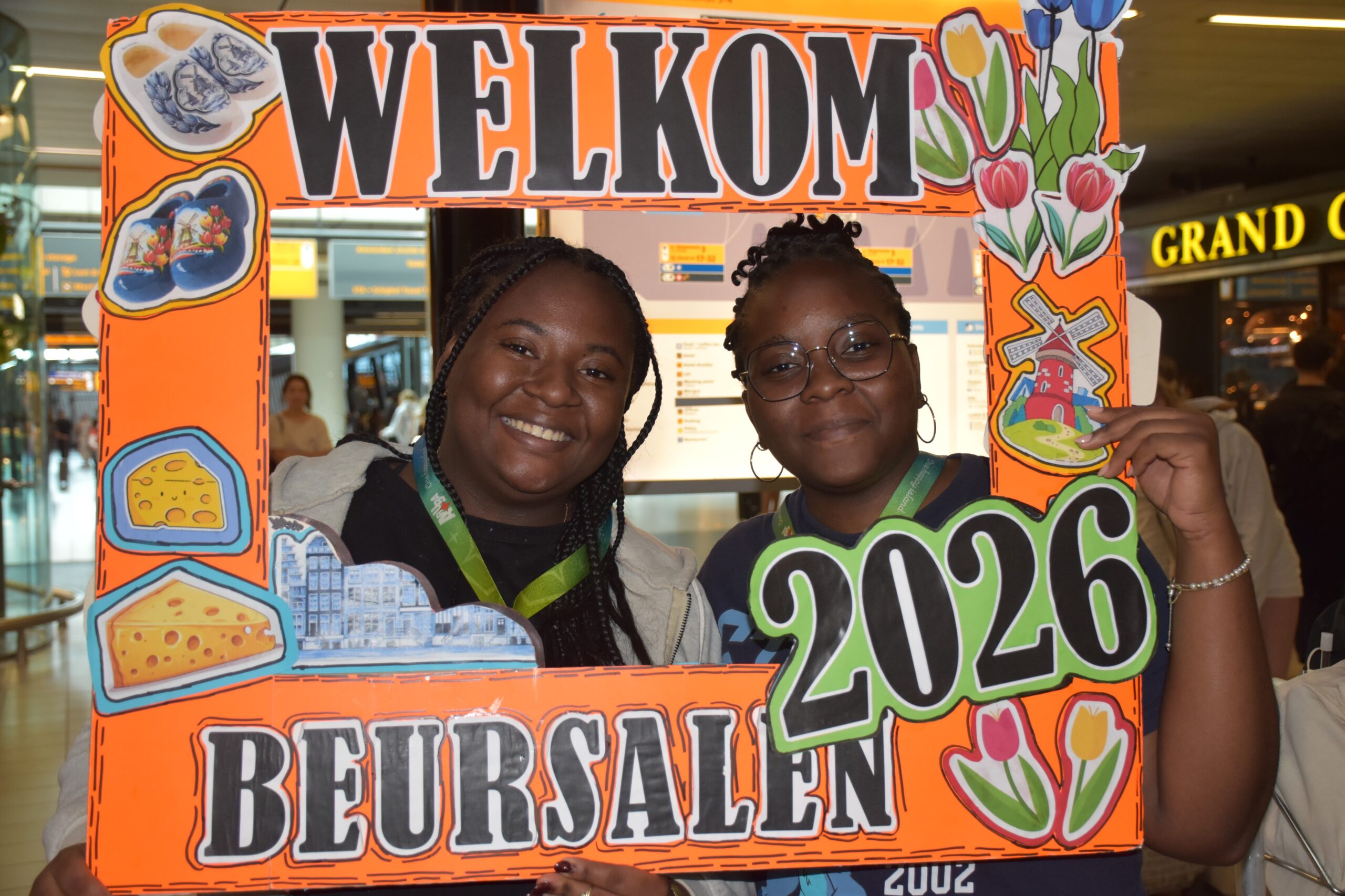 Two smiling people pose behind a bright orange welcome frame in a busy indoor space, like an airport or mall.