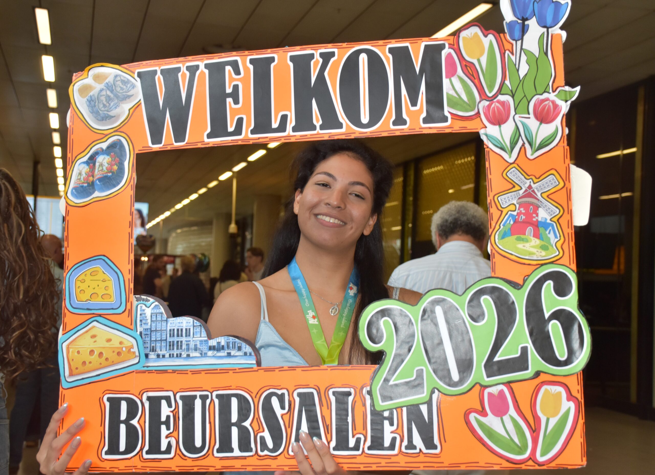 Smiling woman posing behind a large orange decorative frame that says 'WELCOME' with colorful flowers and food illustrations indoors.