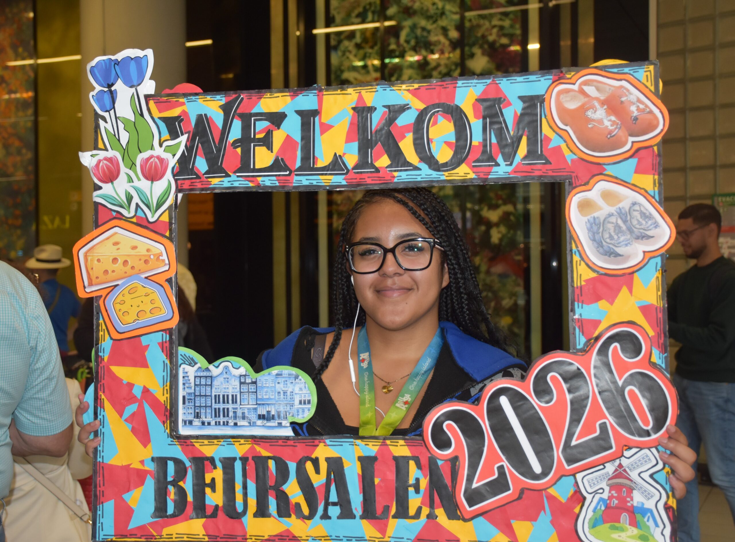 Young woman with braided hair and glasses posing behind a colorful photo-frame prop that says 'WELCOME' with food illustrations, at an event.