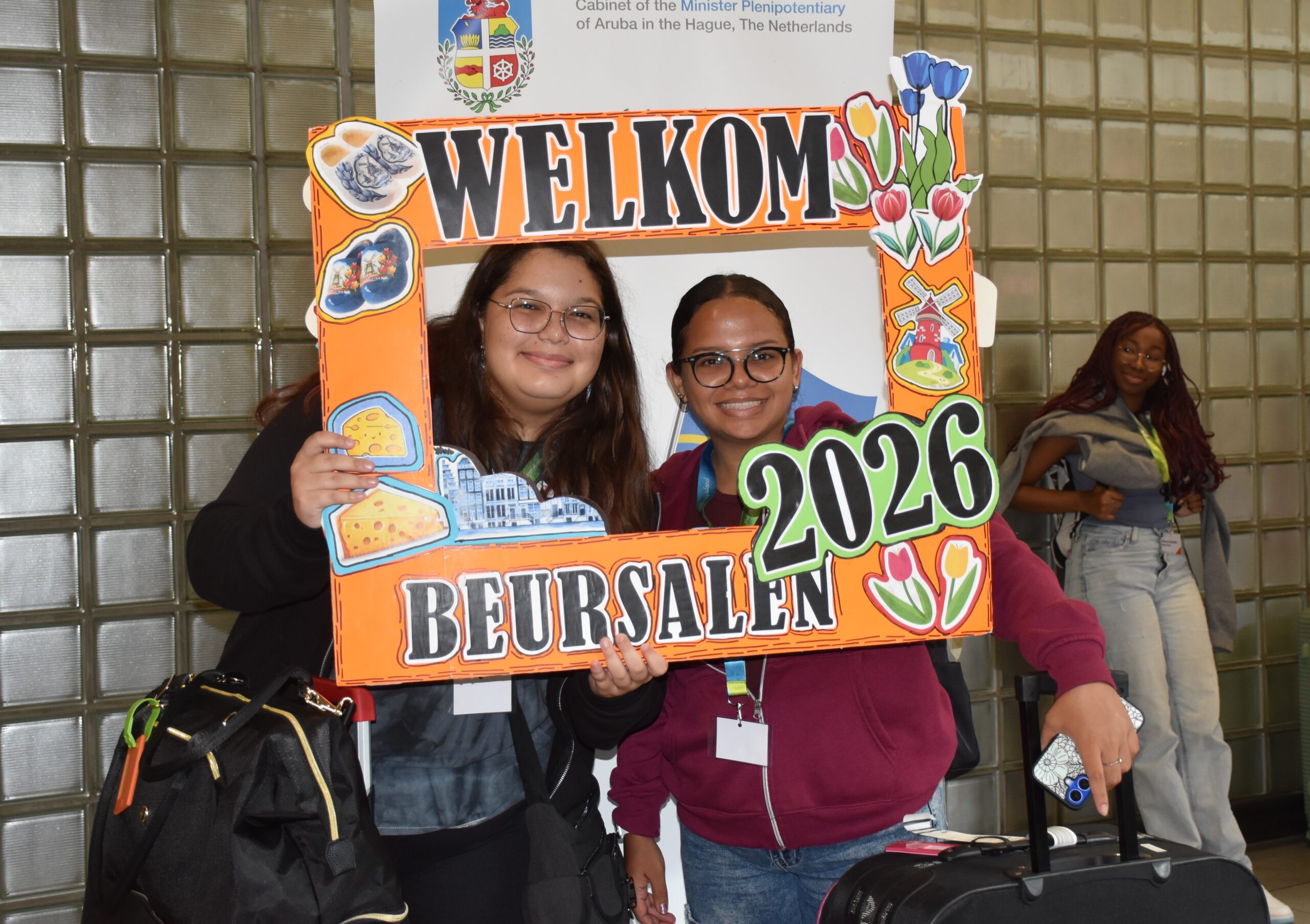 Two smiling young women hold an orange decorative frame that says 'WELCOME 2026' at a travel or welcome event.
