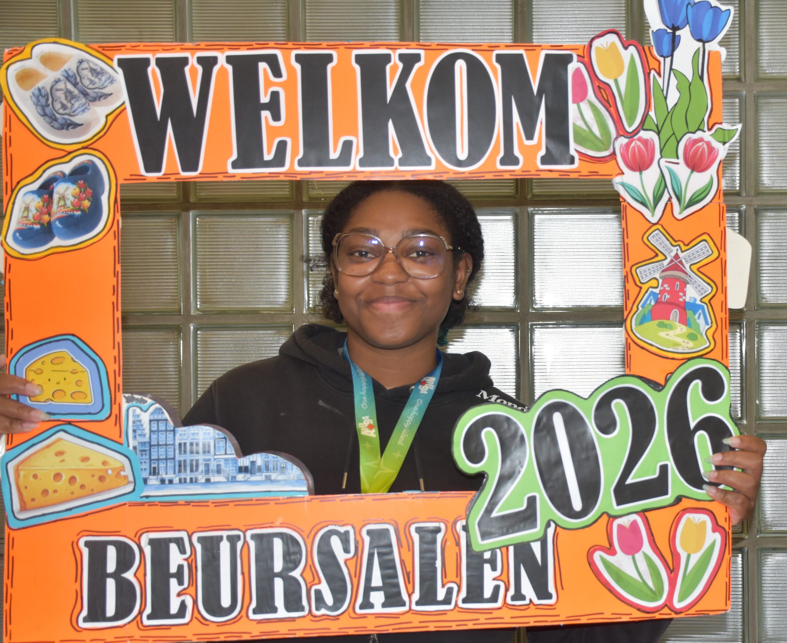 Smiling person with glasses holds an orange decorative 'WELCOME' sign with flowers and colorful illustrations on the sides.