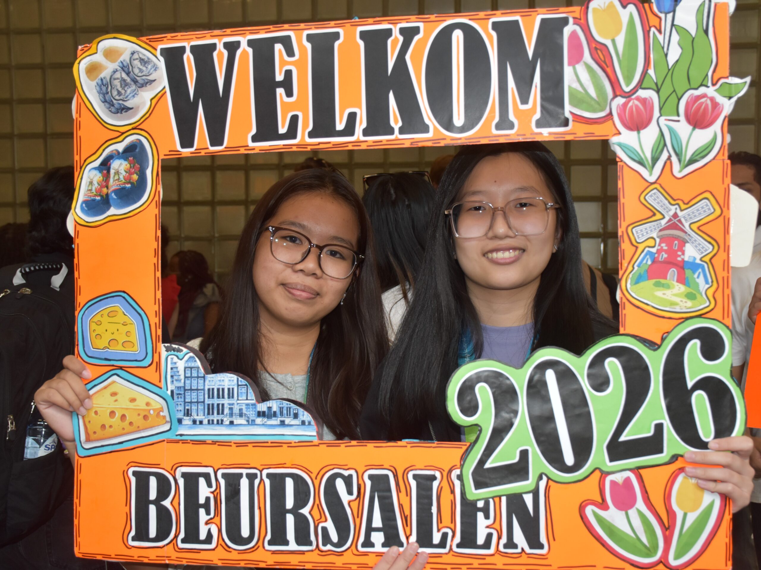 Two young women smile behind a bright orange, cutout welcome frame decorated with food and flower illustrations.