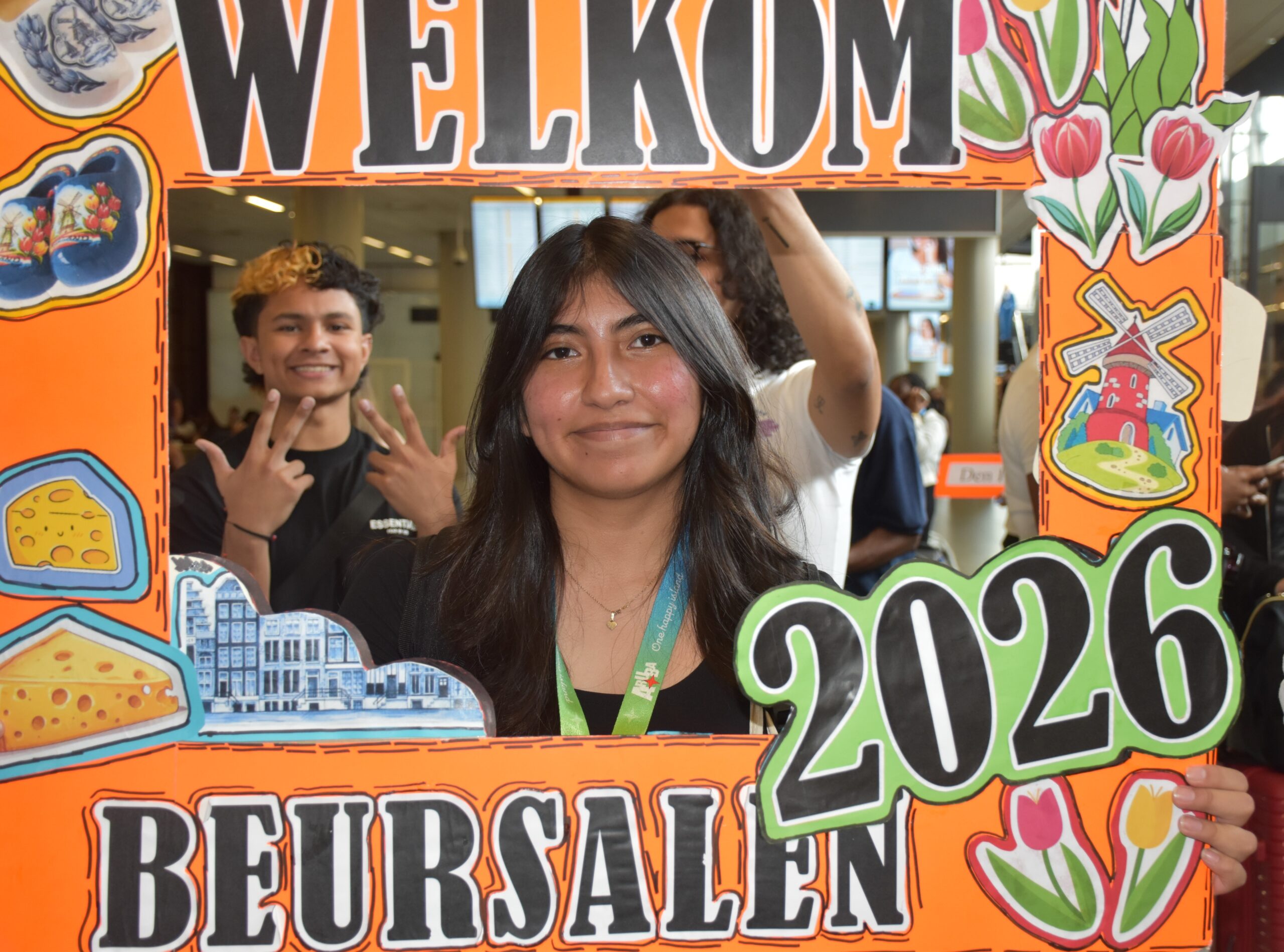 Smiling young people posing behind a bright orange illustrated photo-frame sign with the word 'WEEK' at the top and food drawings around the frame.