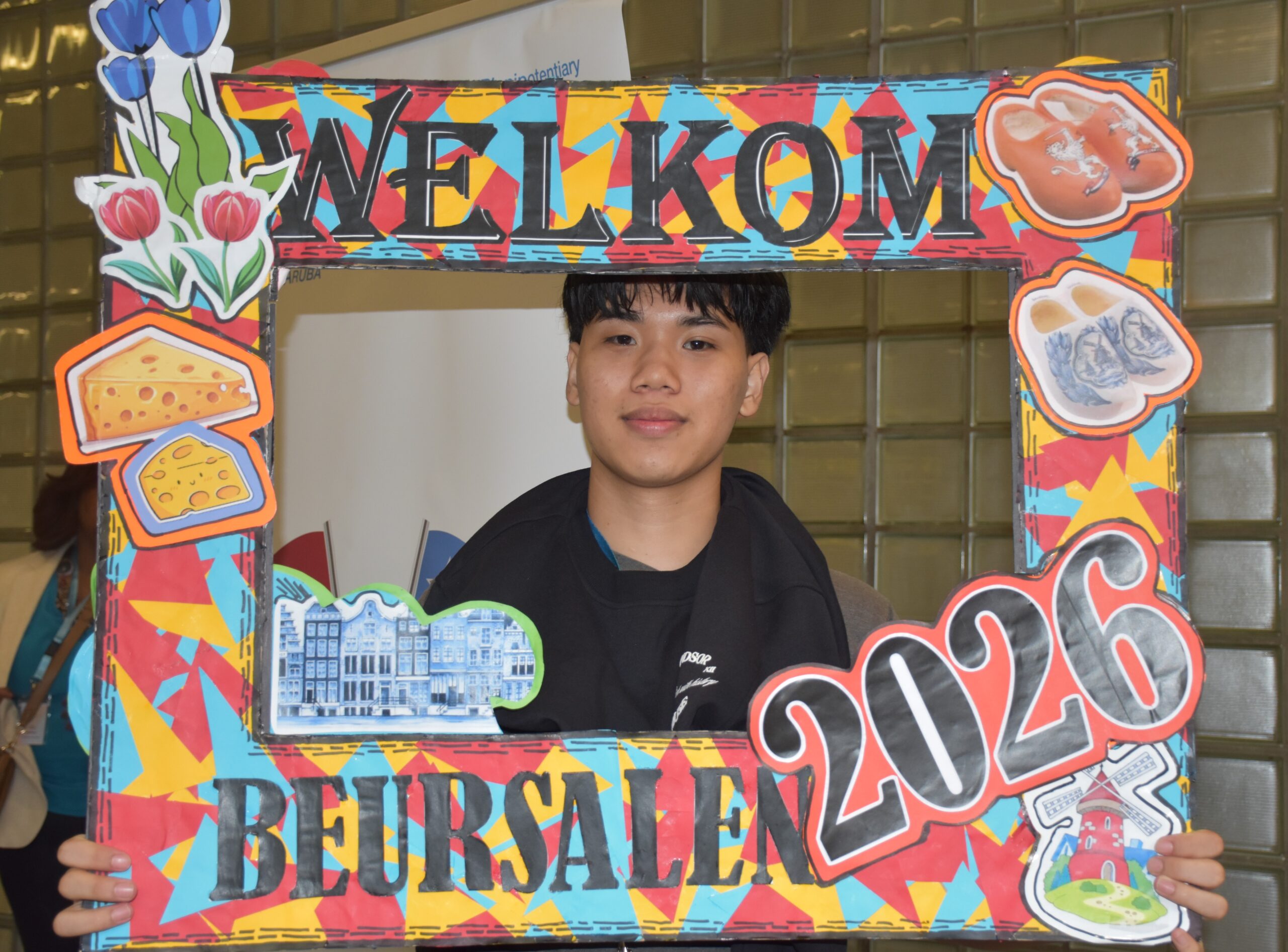 Young man posing behind a bright, decorative frame that reads 'WELCOME' with cartoon food illustrations around it (cheese, bread).