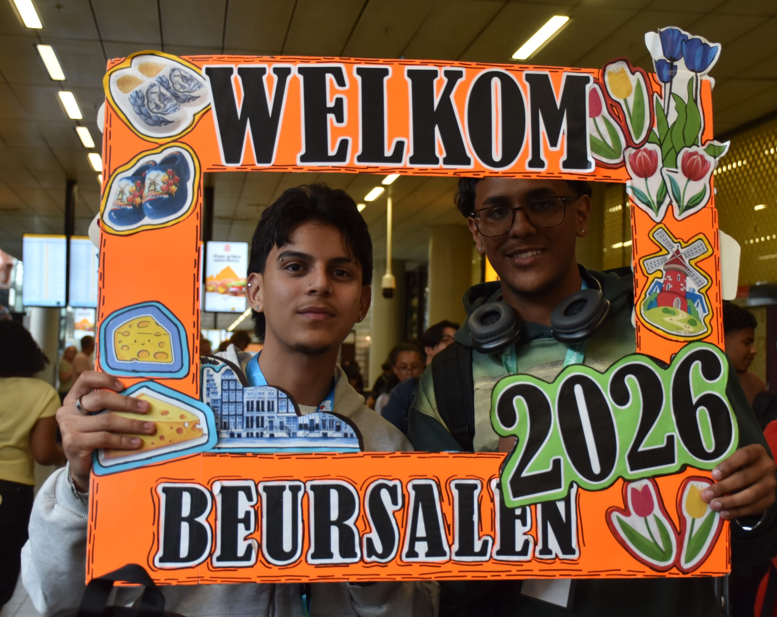 Two young men pose behind a bright orange 'WELCOME' photo prop decorated with food illustrations and flowers at a busy indoor event.