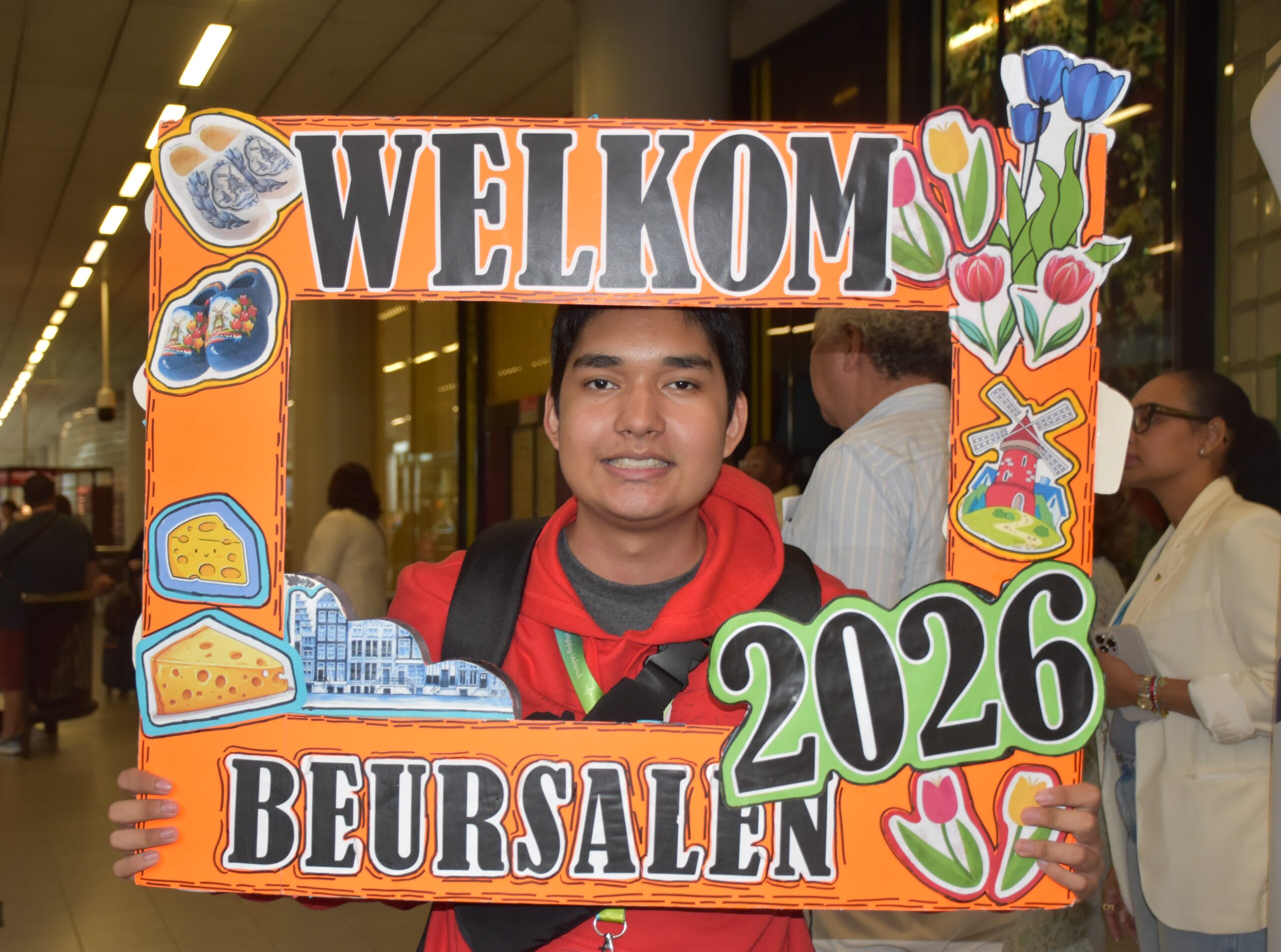 Young man in a red hoodie holds a bright orange “WELCOME” sign decorated with flowers and food illustrations in a mall.