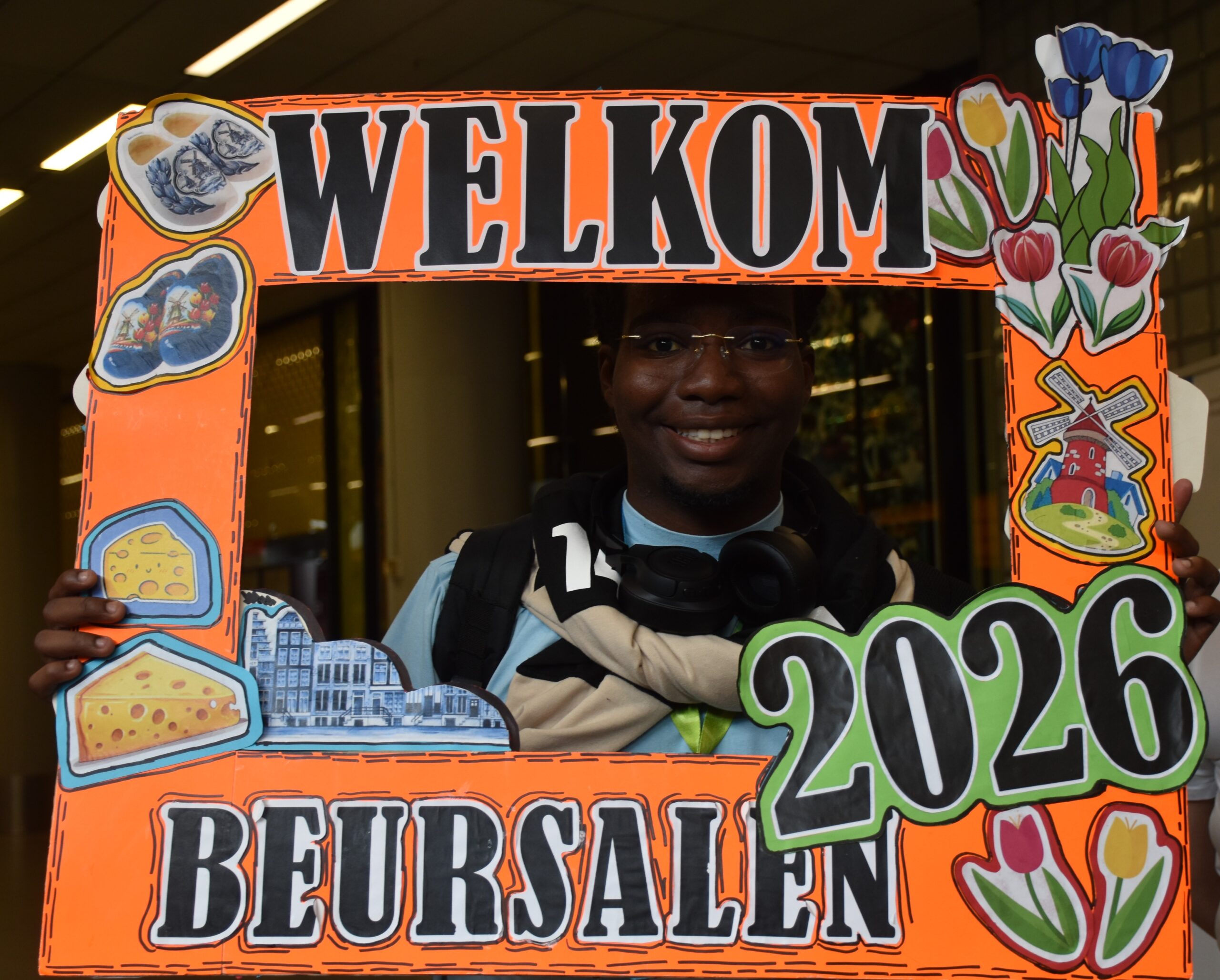 Person smiling behind a bright orange welcome sign framed with cartoon cheese, flowers, and buildings.