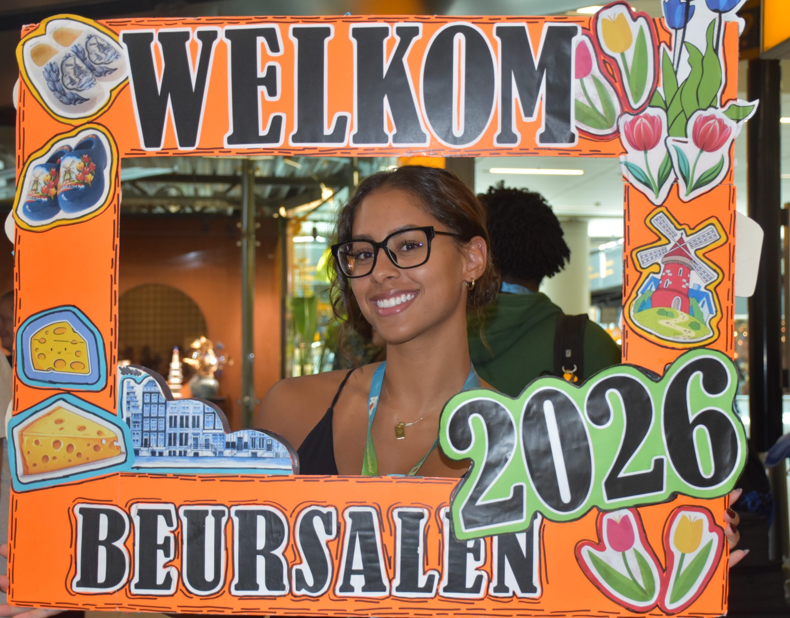Smiling woman with glasses behind a bright orange Welcome sign decorated with flowers, cheese, and buildings graphic elements.