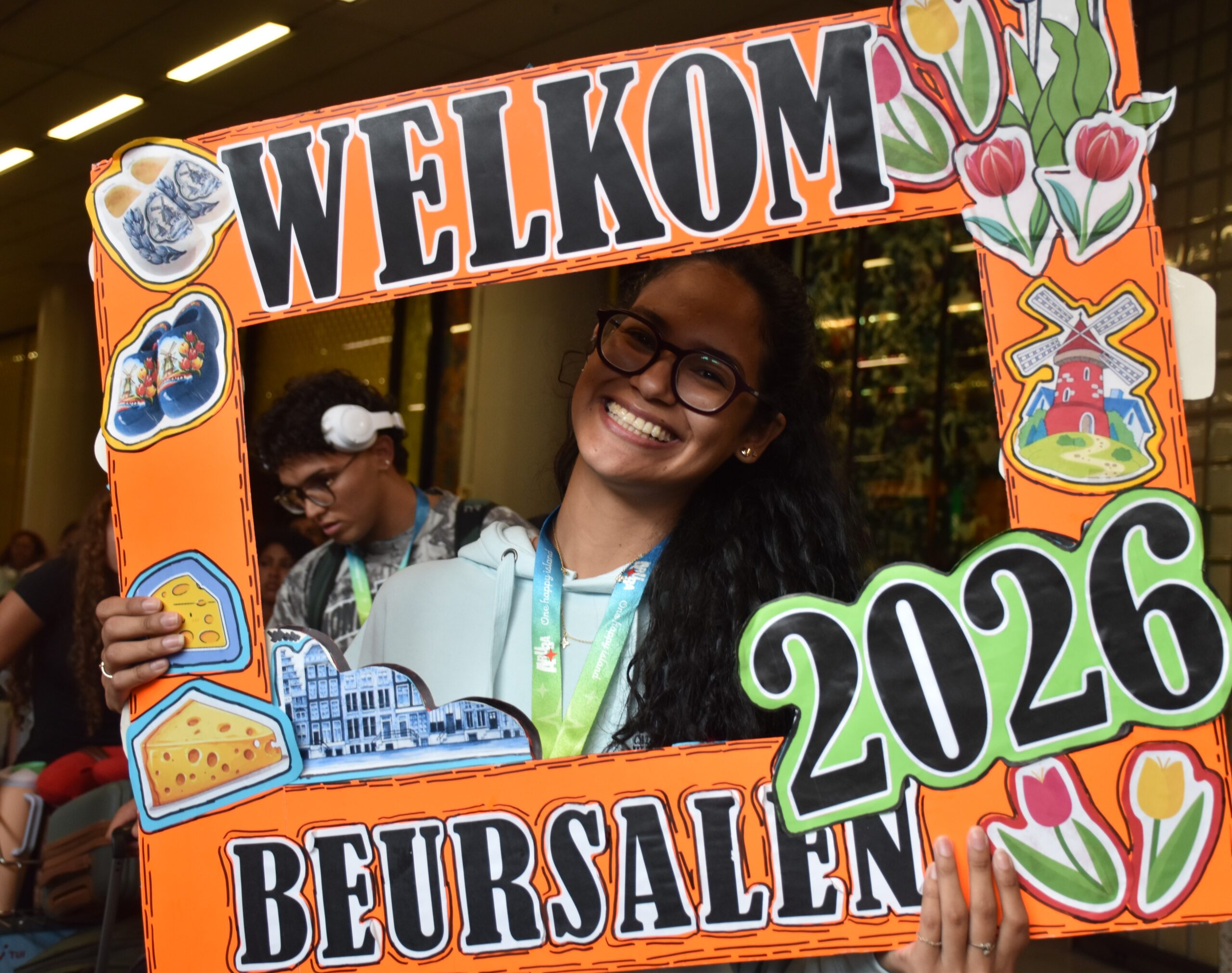 Smiling woman with glasses holds a bright orange decorative welcome sign at an event.
