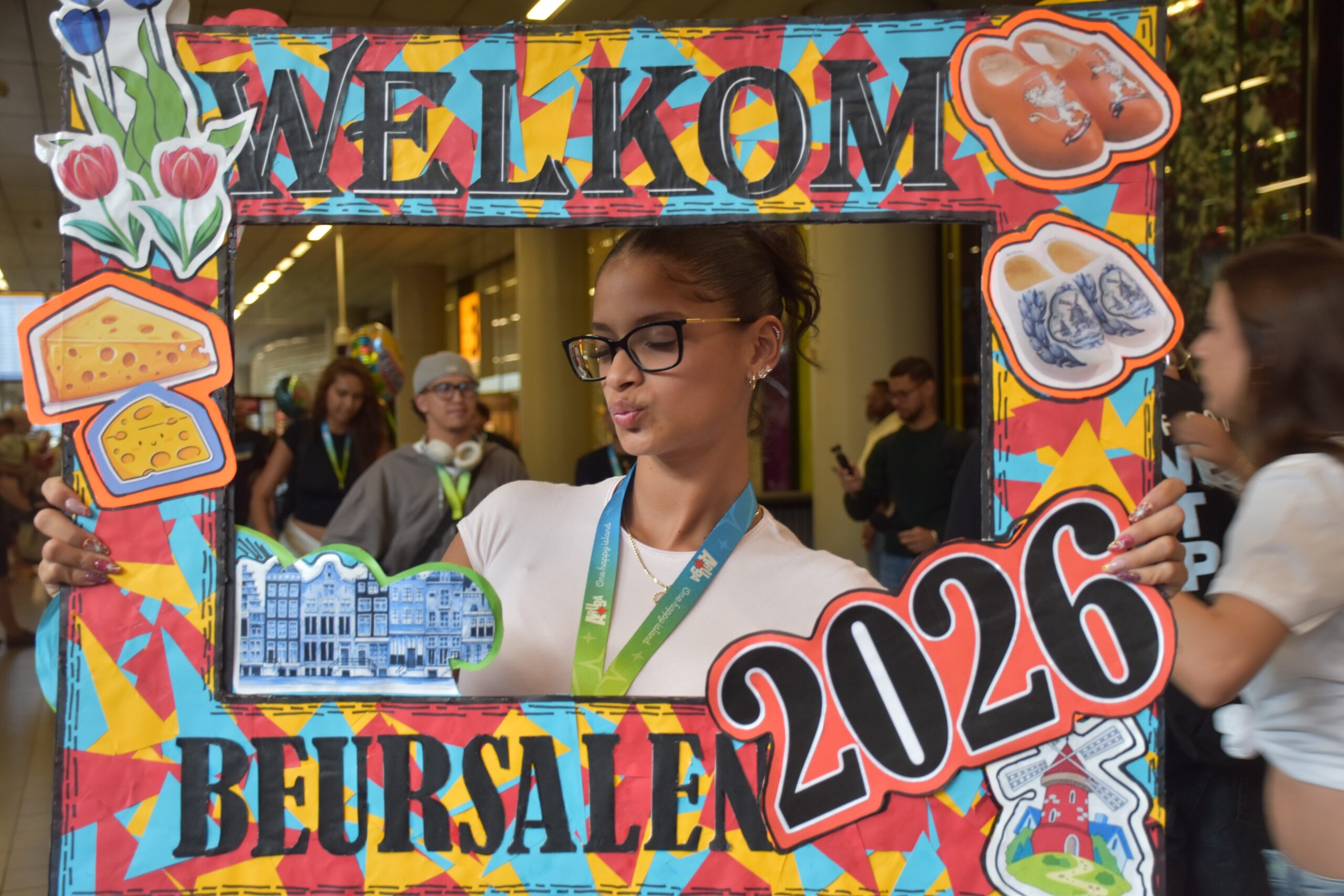 Woman with glasses posing behind a bright, collage-style welcome frame with cheese and other food cutouts; people in a indoor event in the background.