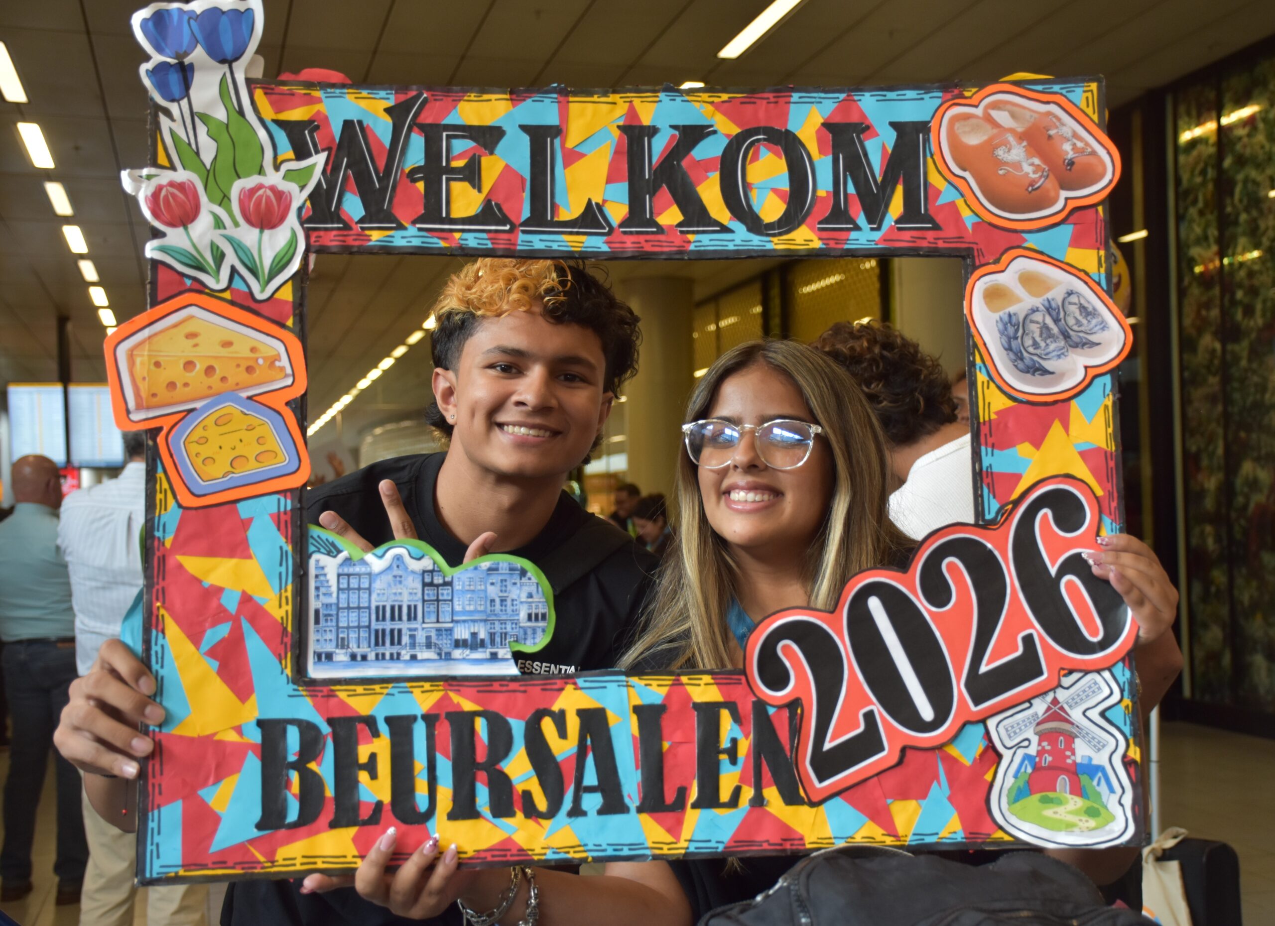 Two smiling teenagers pose behind a colorful, graffiti-style welcome frame with food and flower decorations.