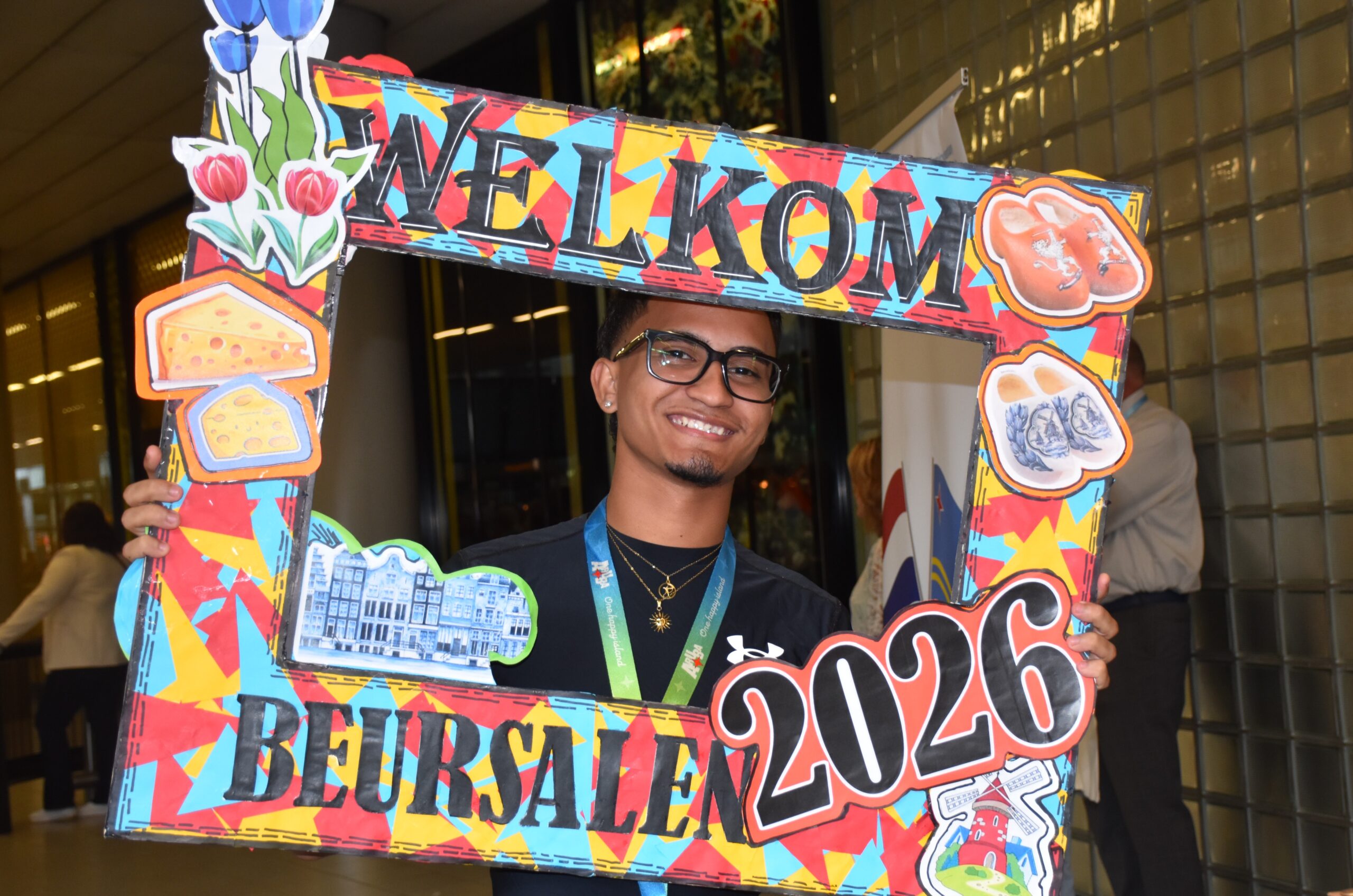 Smiling young man holding a large colorful frame decorated with food images (cheese, bread) at an indoor event.