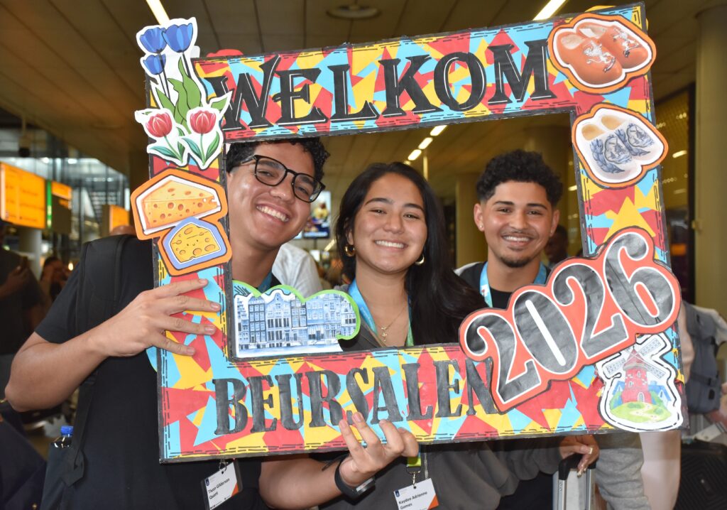 Three smiling young adults posing behind a bright, decorative frame that says WELCOME at a crowded event.