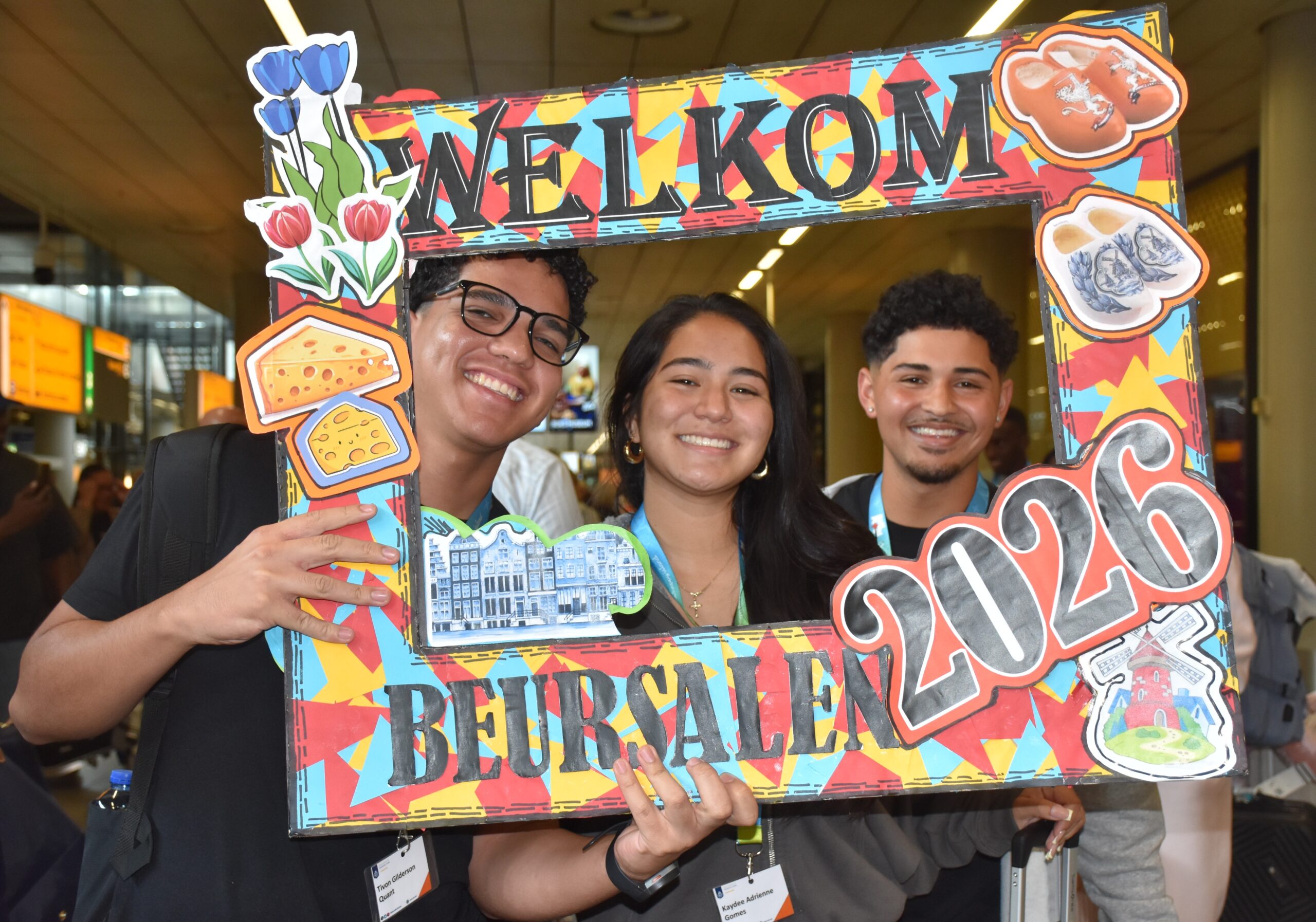 Three smiling young adults posing behind a bright, decorative frame that says WELCOME at a crowded event.