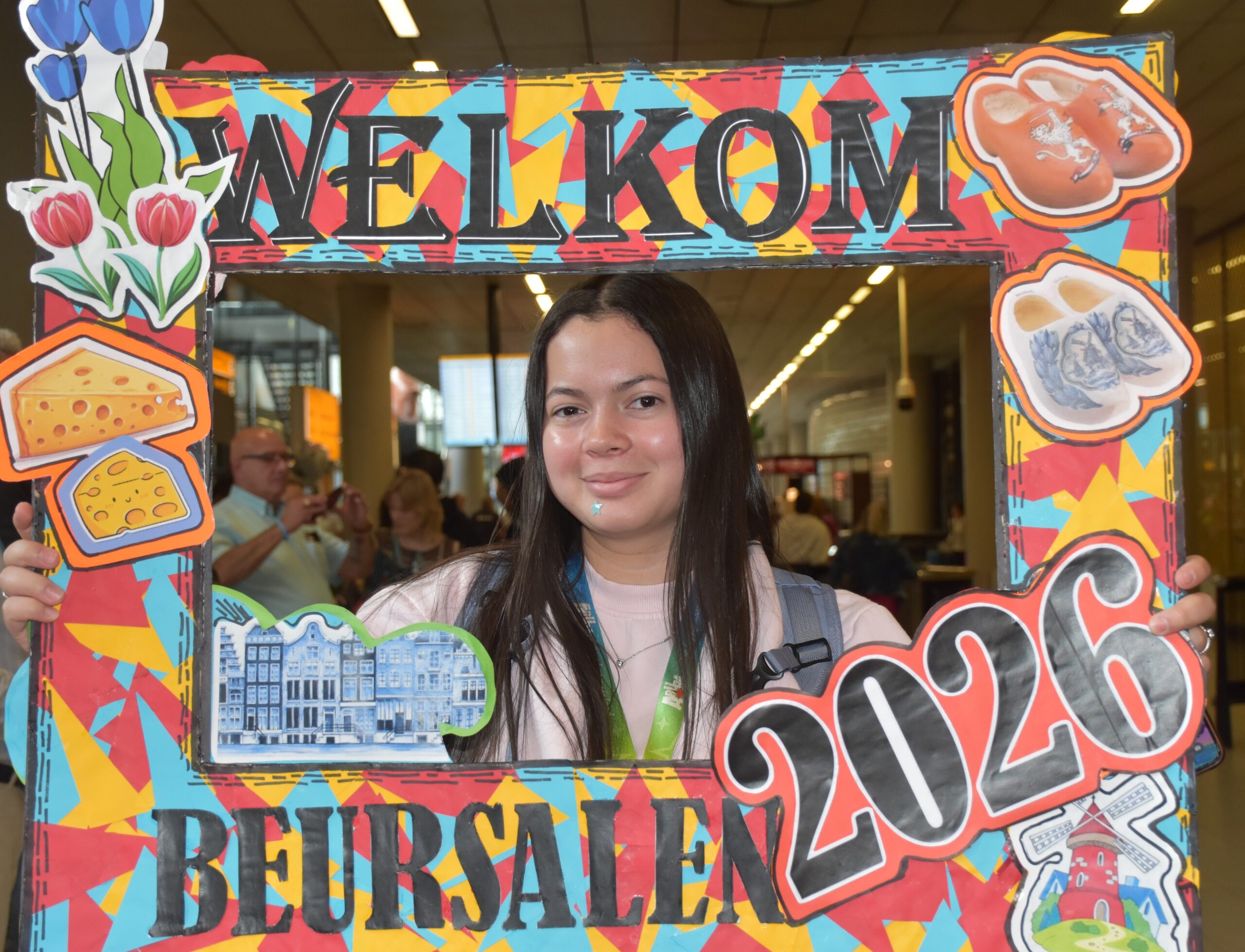 Woman posing behind a bright, decorative 'WELCOME' frame with food illustrations at an event hall null background blur of people behind her.