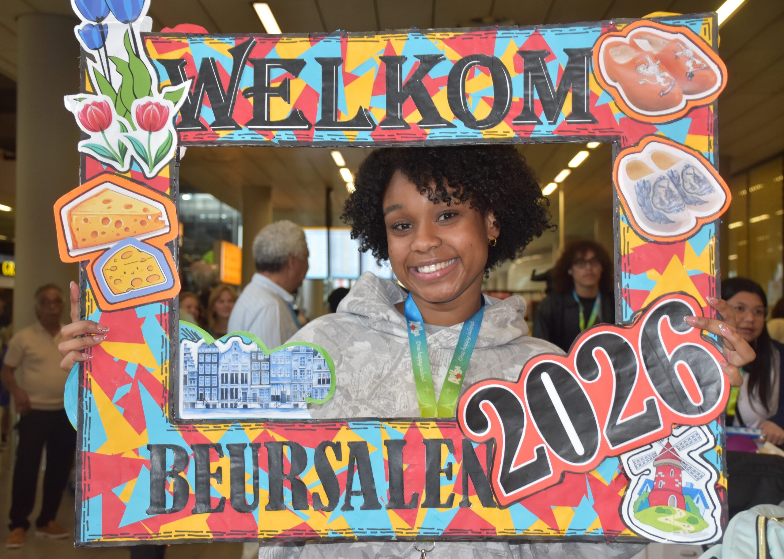 Smiling young woman posing behind a colorful, decorative frame with food stickers (cheese, bread) at an event.
