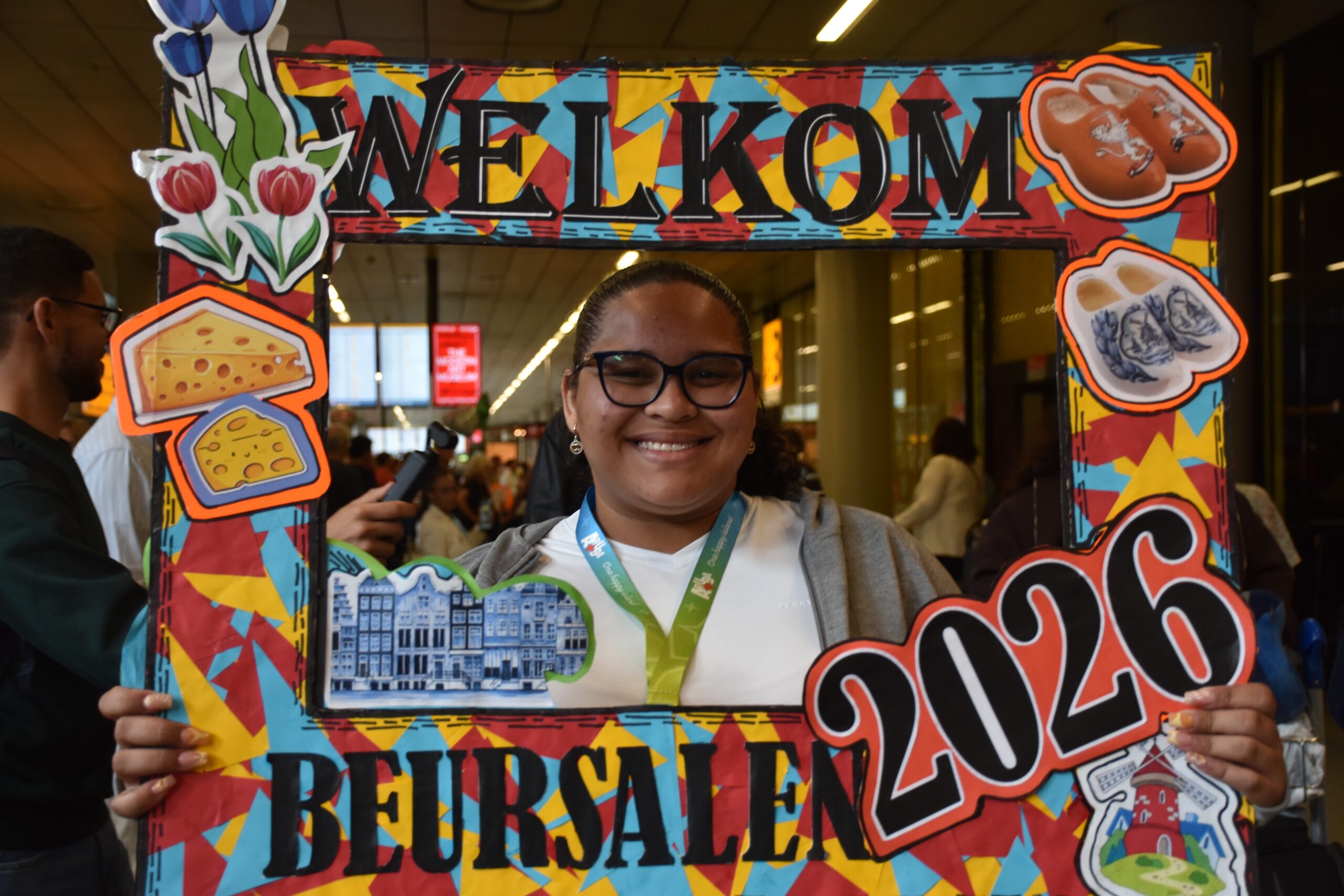 Smiling person poses behind a colorful, cut‑out welcome frame with food-themed stickers at an event.