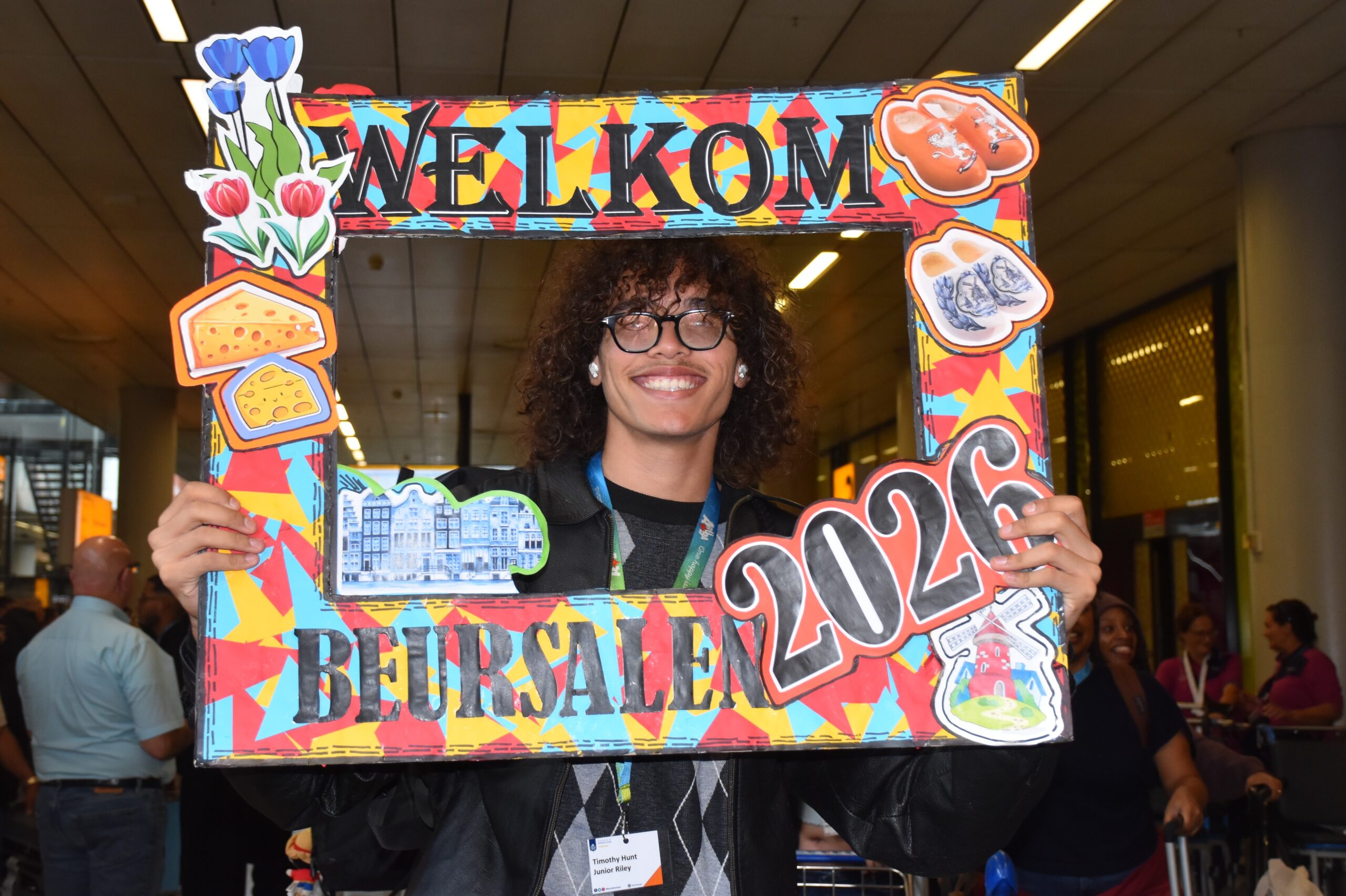 Man with curly hair and glasses smiles while holding a bright, collage-style Welcome photo frame at an event.