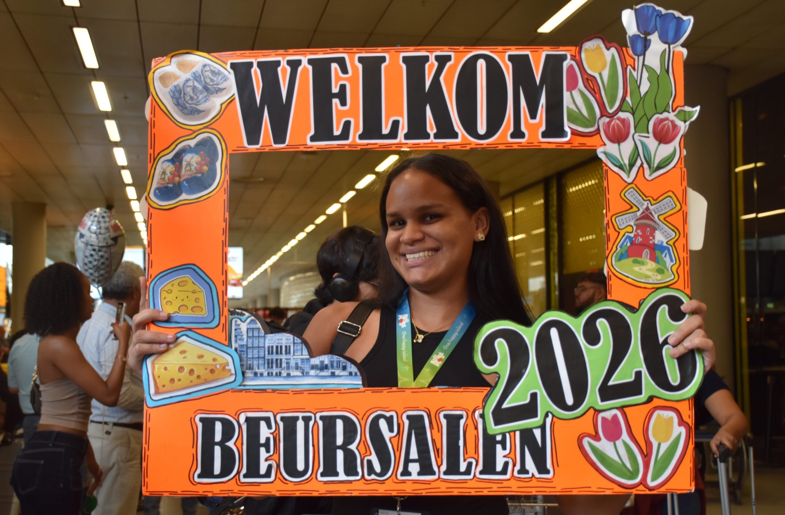 Smiling woman posing behind a bright orange decorative frame that says 'WELCOME' with flowers and food stickers at an indoor event.