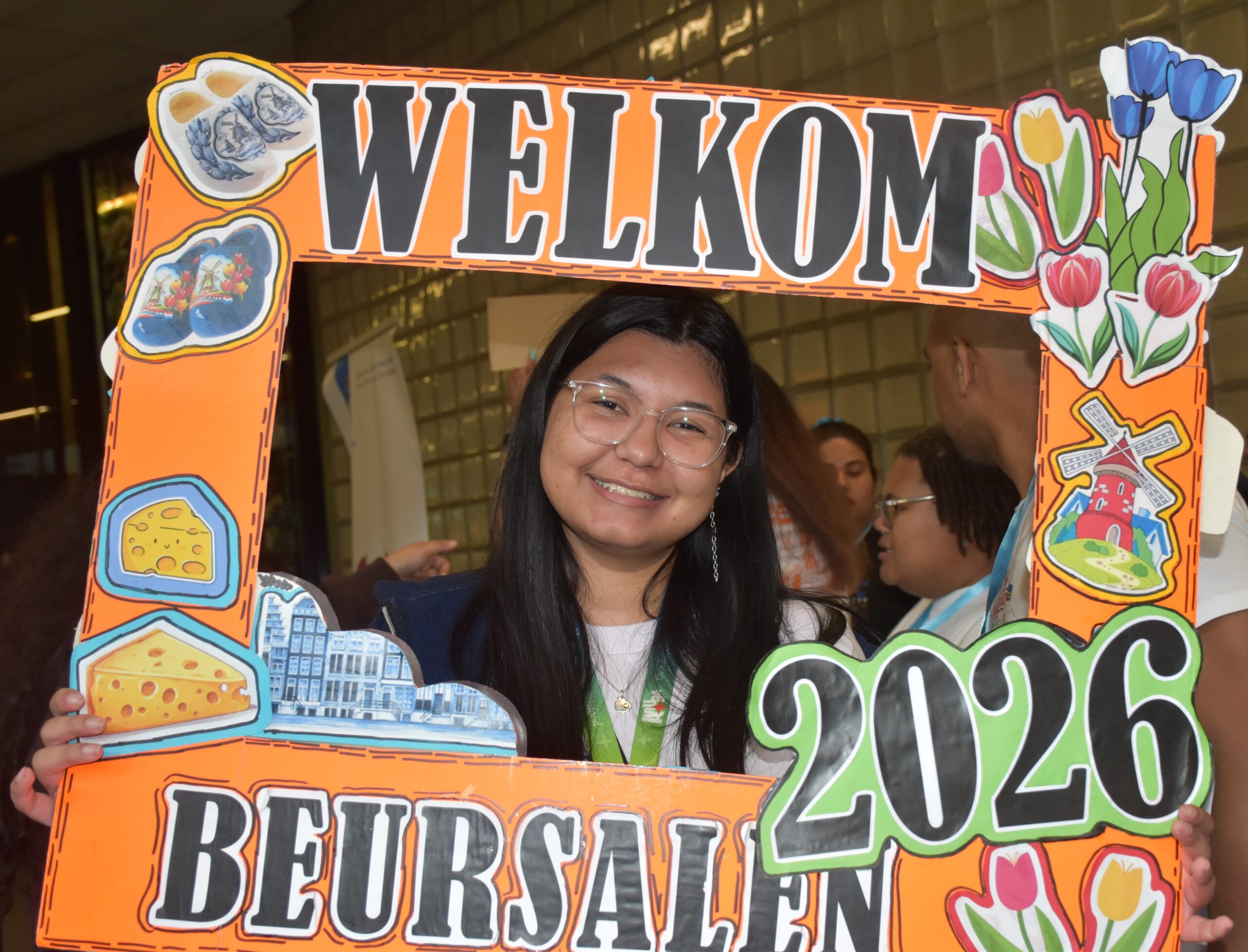 Person smiling behind a bright orange, cut-out “WELCOME” sign with drawings of food, flowers, and city images.