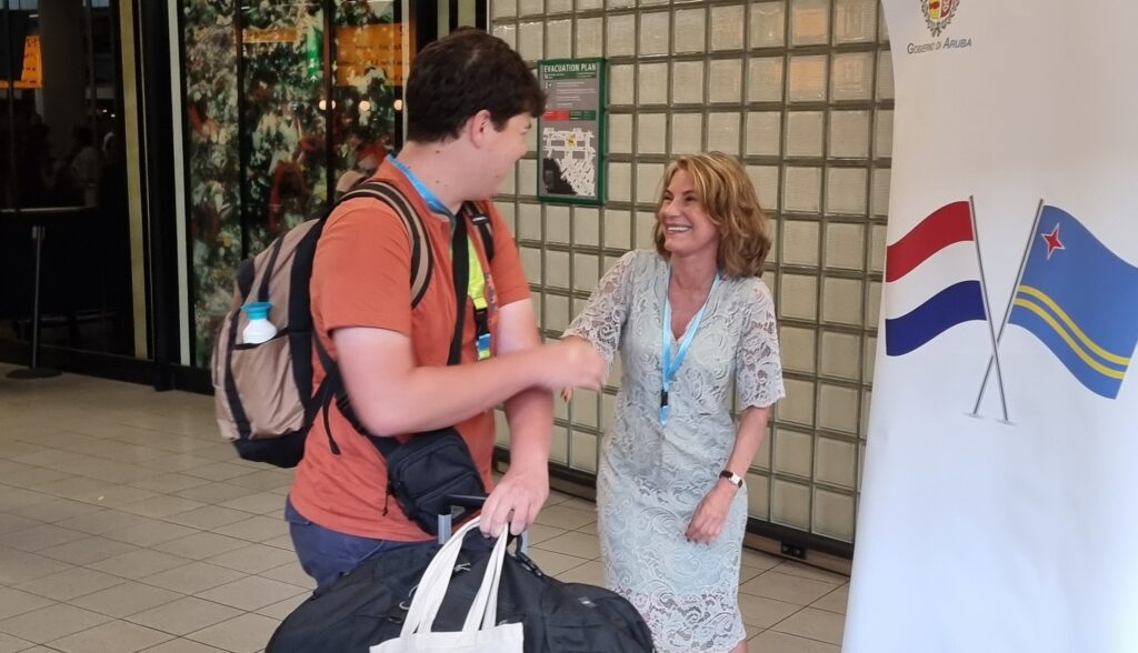 Man in orange shirt shaking hands with smiling woman in lace dress, carrying a large bag, near a government banner with Aruba flags.
