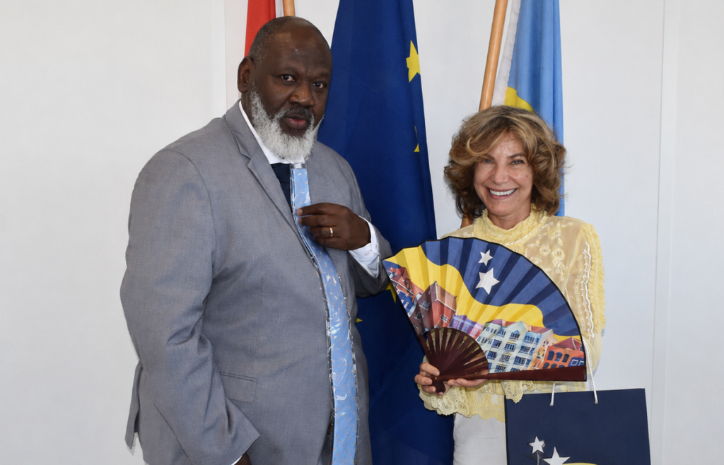 Two people pose for a photo: a man in a gray suit and a smiling woman in a pale yellow blouse holding a decorative fan, with flags in the background.
